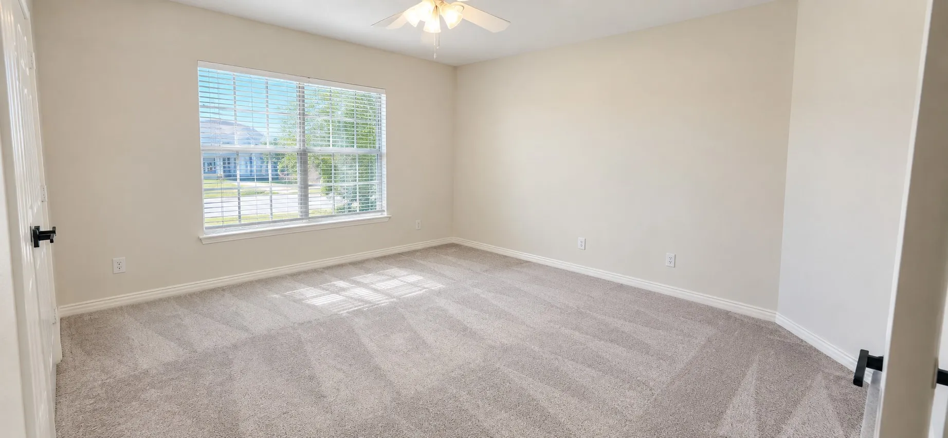Spacious room featuring neutral carpeting, a large window with blinds, and an overhead ceiling fan