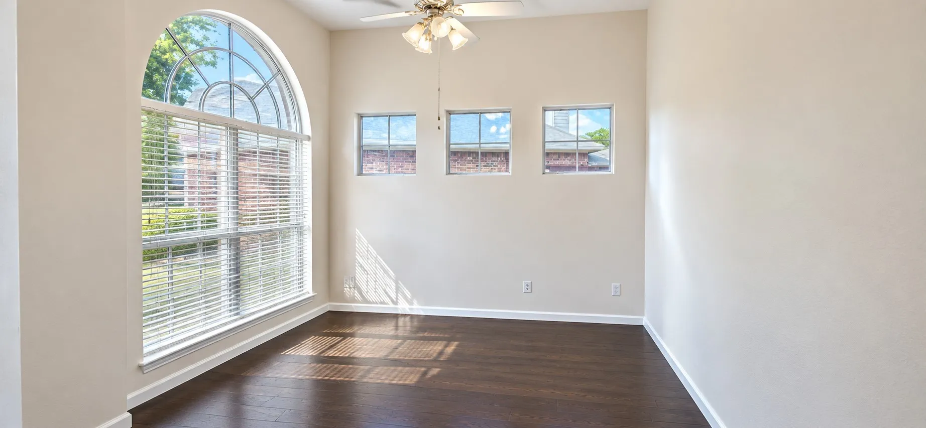 Arched window with gridded pane, three rectangular windows, dark wood-finish flooring, and a ceiling fan with integrated lighting. could be used as office or bedroom.