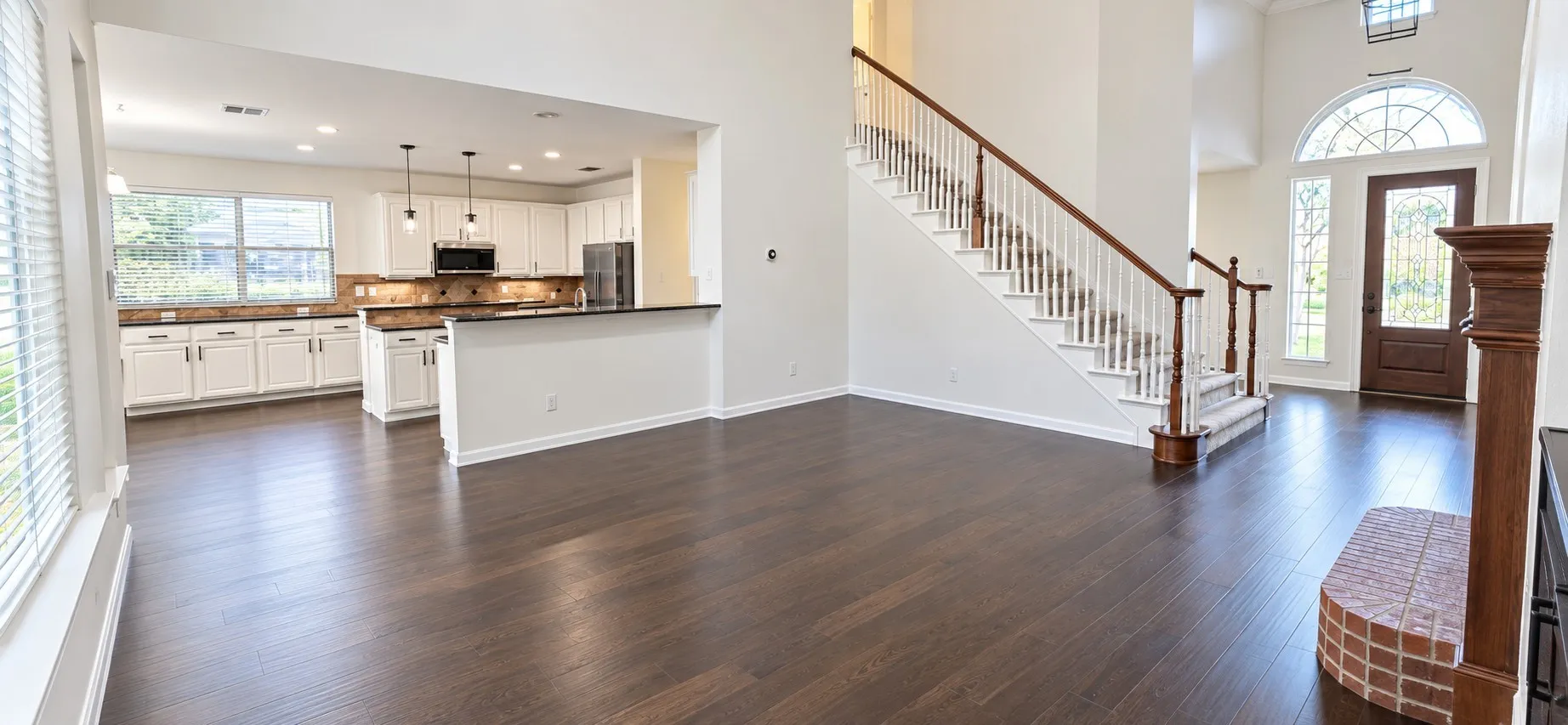 Spacious open-concept living area featuring dark wood-finish flooring, a two-story foyer, a grand staircase with wood railing, high ceilings and fireplae with shelving on both sides