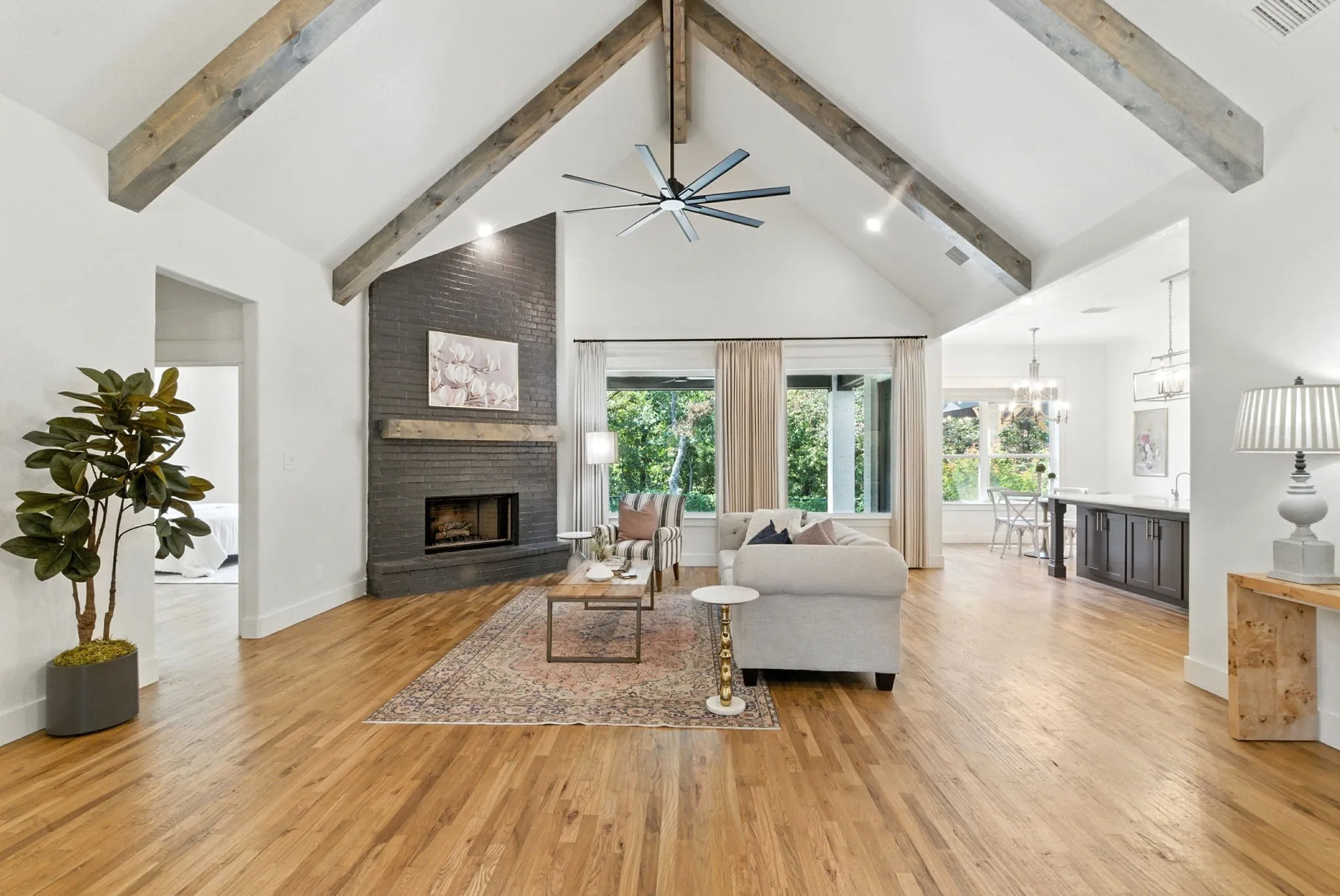 Vaulted ceilings with exposed wood beams, a dark brick fireplace with a wood mantle, large windows with natural lighting, and wood-finish flooring