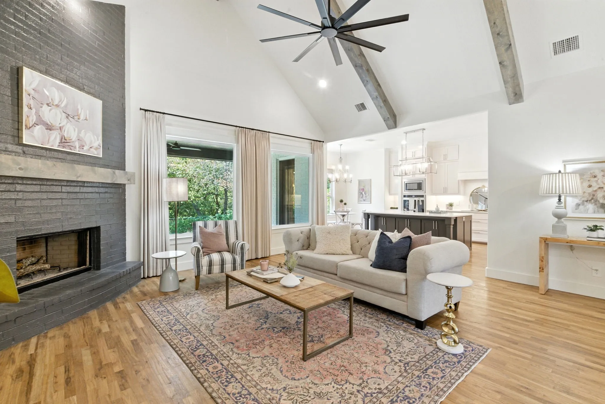 Vaulted ceiling with exposed wood beams, wood-finish flooring, floor-to-ceiling windows, brick fireplace with wood mantel, and kitchen with central island