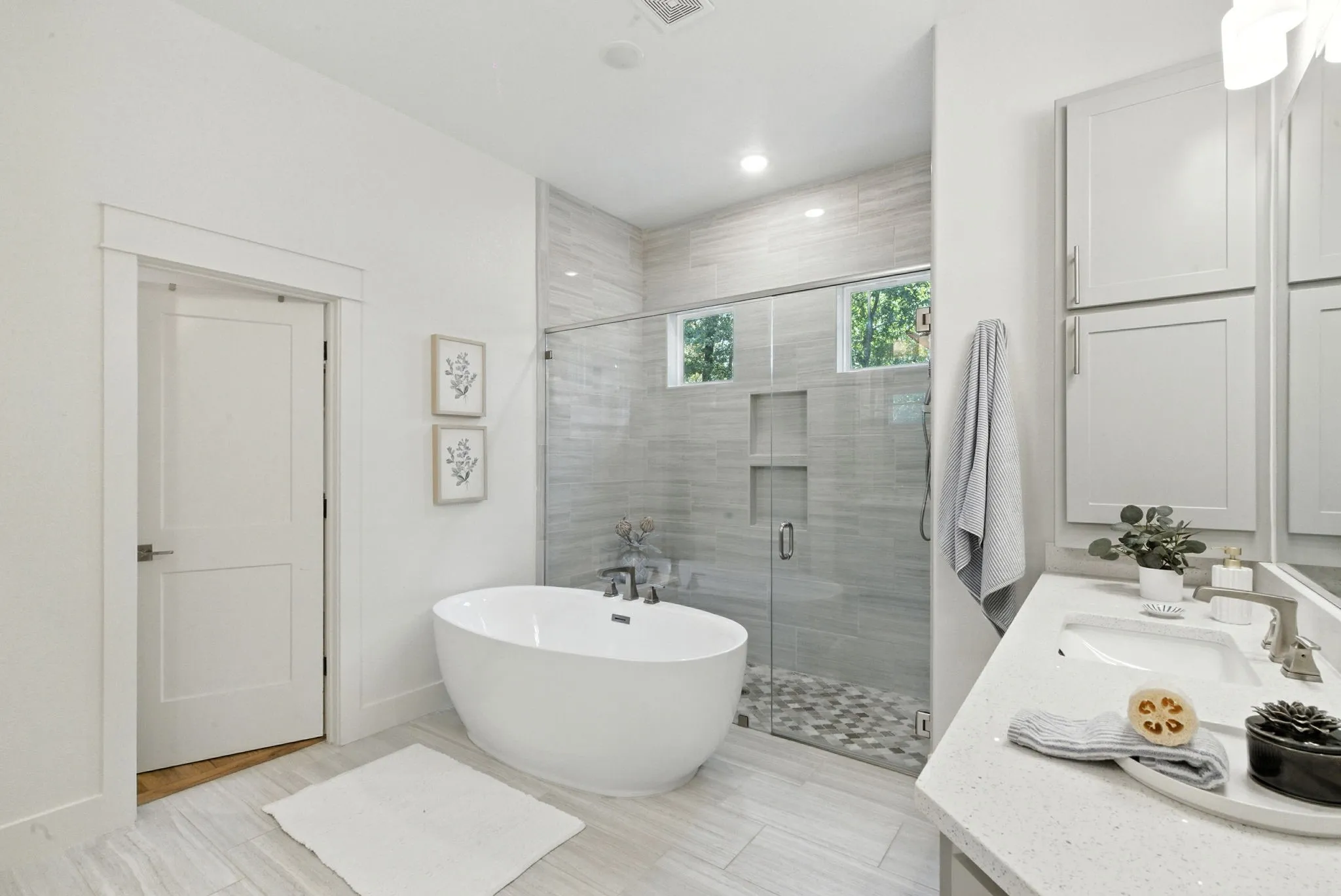 Freestanding soaking tub, glass-enclosed shower with pebble flooring and tiled niche, light wood-finish flooring, dual under-mount sinks, and light-toned cabinetry
