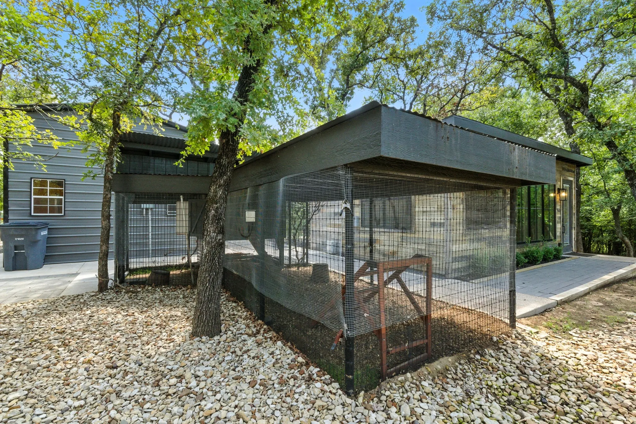 Exterior featuring corrugated metal siding, an outdoor enclosure with metal caging, established trees, a concrete walkway, and a gravel ground cover