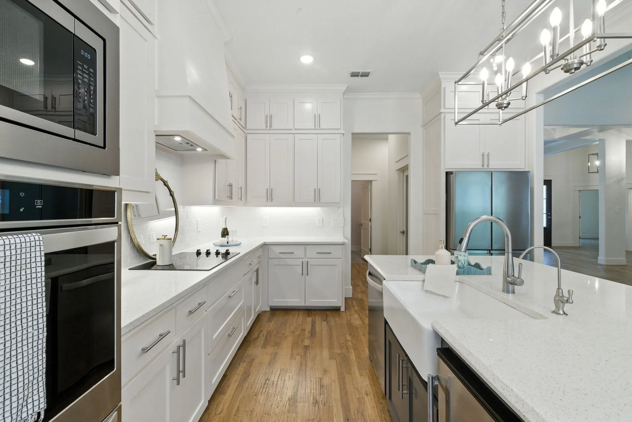 White cabinetry with under-cabinet lighting, stainless steel appliances, wood-finish flooring, a kitchen island with a farmhouse sink, and contemporary light fixtures