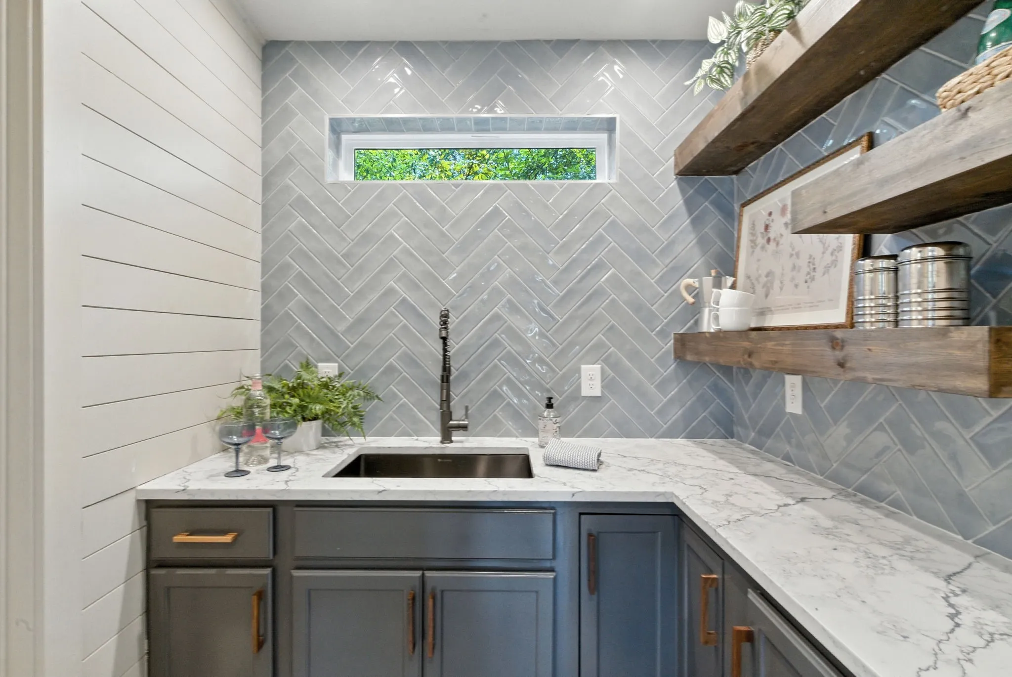White shiplap wall paneling, herringbone tile backsplash, white countertop, dark-finished cabinetry, and wood-finish open shelving