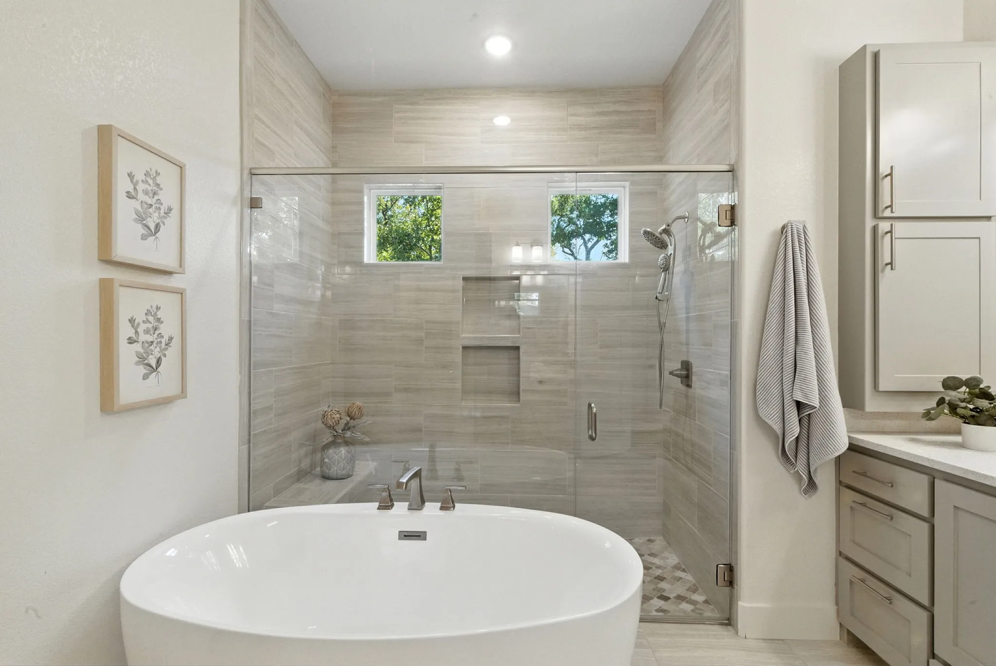 Freestanding soaking tub with brushed nickel fixtures, glass-enclosed shower with tiled walls and built-in niches, dual windows, and recessed lighting