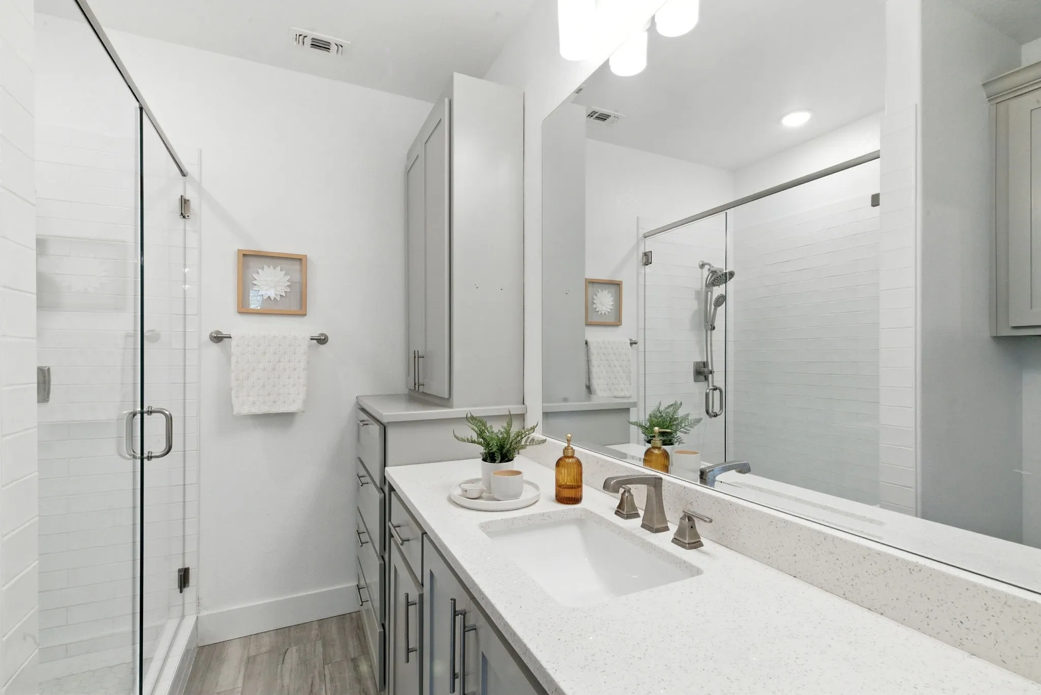 White tile shower enclosure with glass door, vanity with integrated sink, light gray cabinetry, and full-width mirror