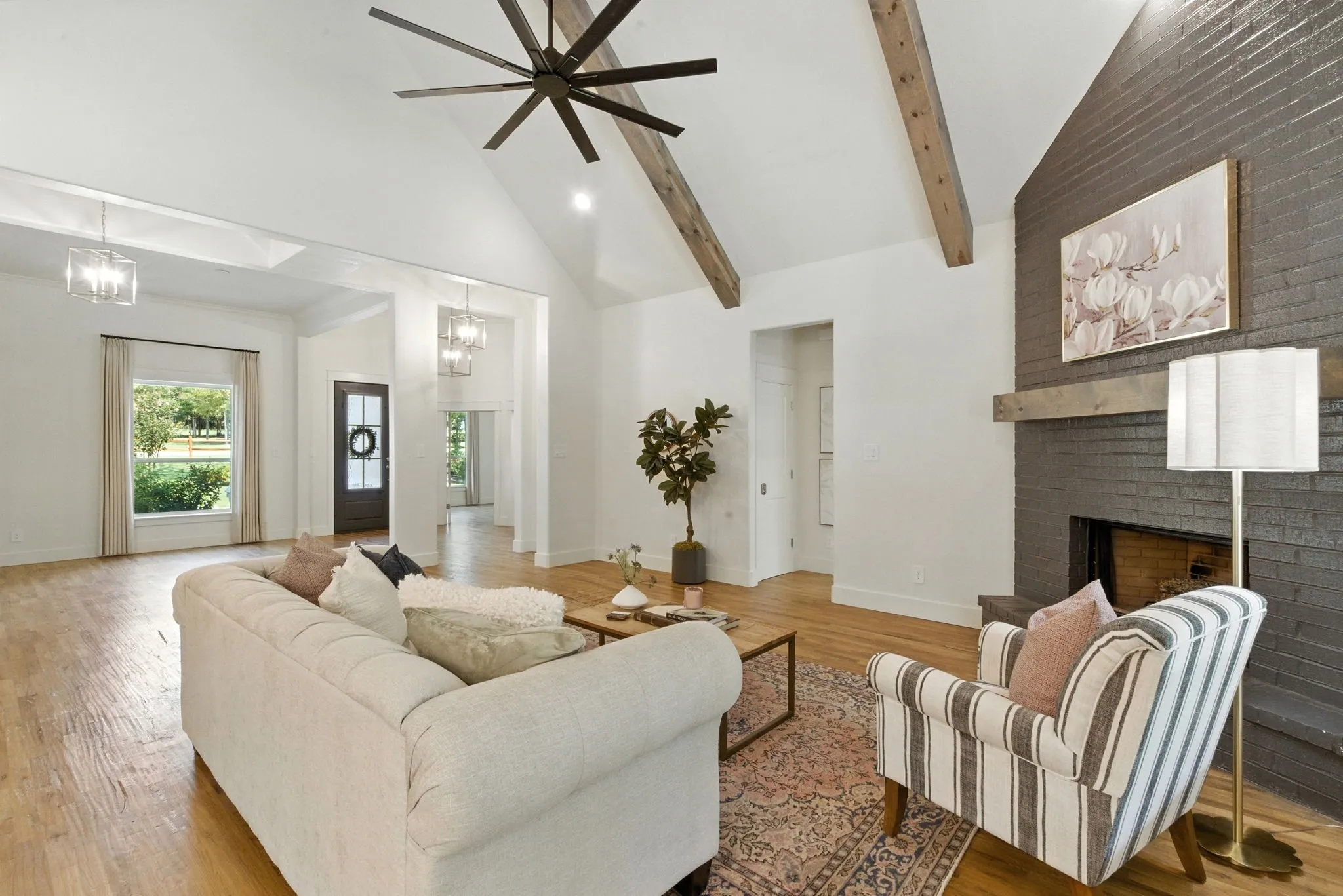 Vaulted ceiling with exposed wood beams, wood-finish flooring, dark brick fireplace with wood mantel, large ceiling fan, and large windows