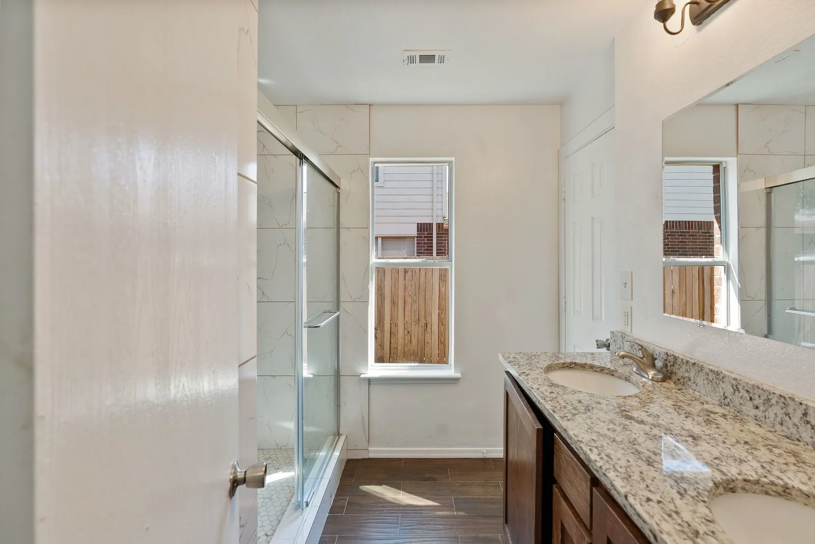 Double vanity with granite countertop, wood-finish flooring, and a glass-enclosed shower featuring white tile surround