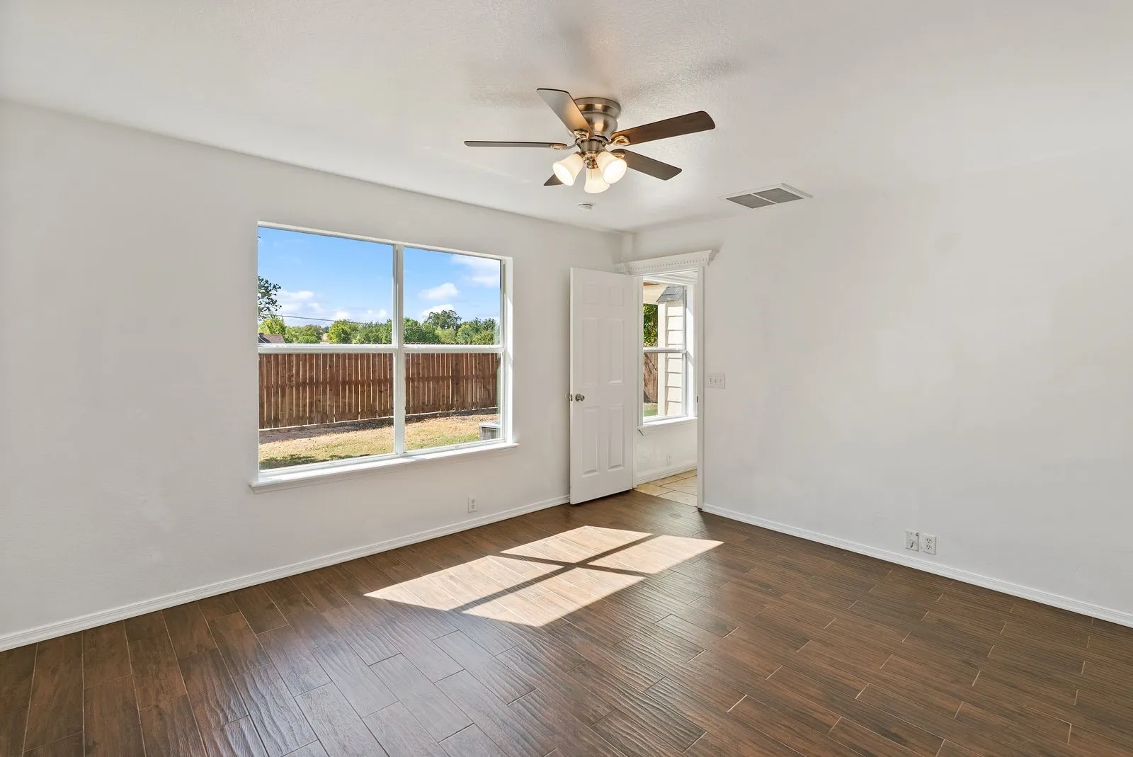 Spacious room featuring wood-finish tile flooring, a ceiling fan with integrated lighting, and a large window overlooking a fenced yard