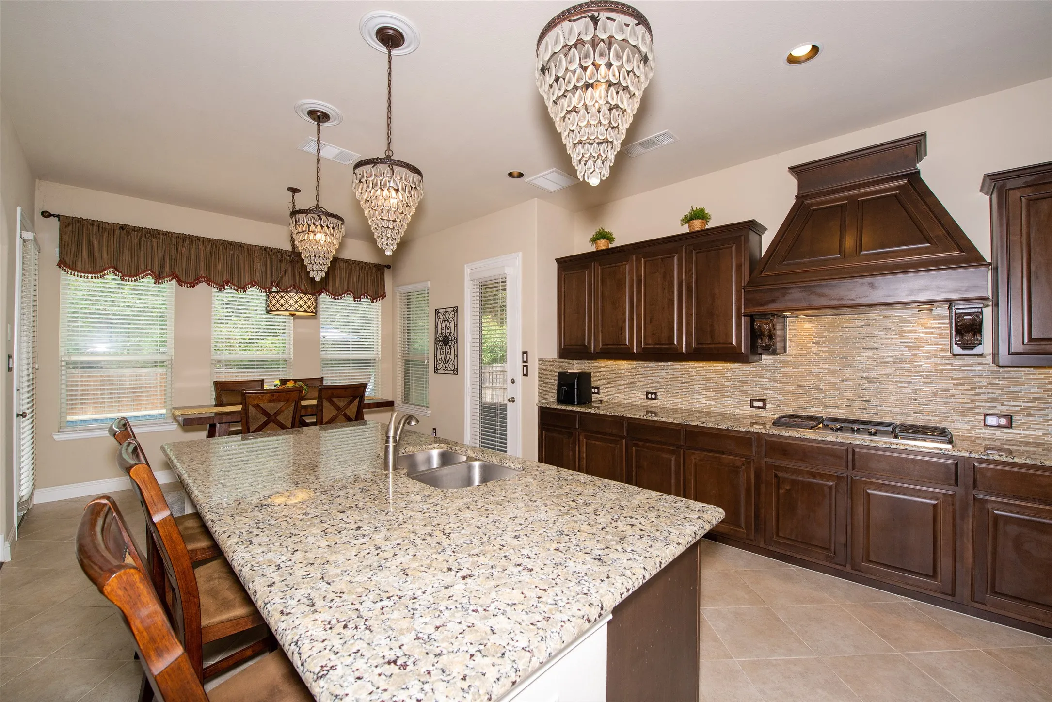 Kitchen featuring dark wood cabinetry, light-toned tiled flooring, a granite-finish island with an undermount double sink, and a tiled backsplash