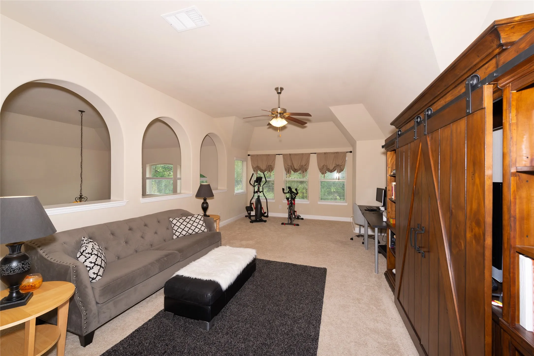 Carpeted space featuring a built-in wood-finish cabinet with barn door hardware, multiple arched openings, a ceiling fan, and multiple windows