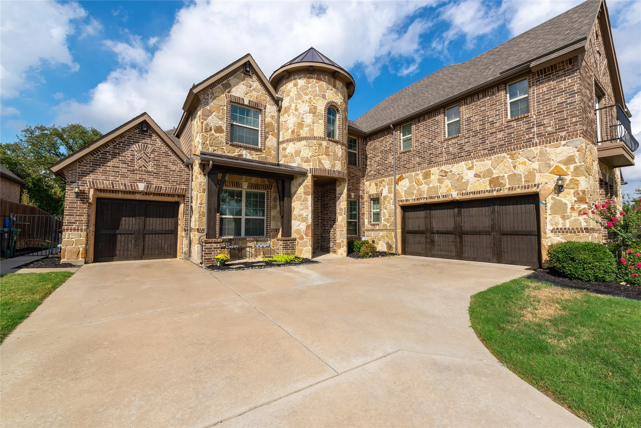 Stone and brick exterior with a turret feature, two attached garages, a second-story balcony, and a concrete driveway