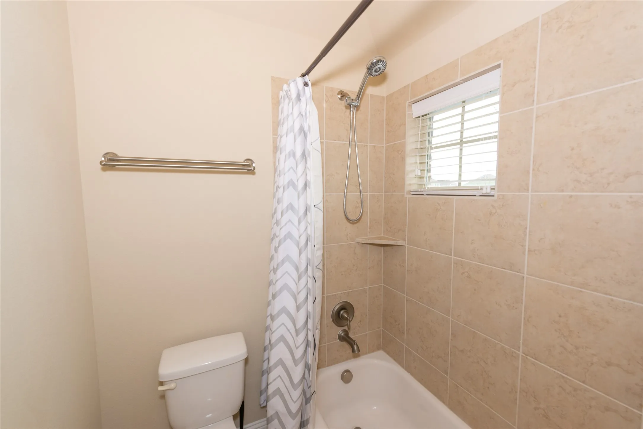 Bathtub with a tiled surround, integrated shower shelf, and a window featuring blinds