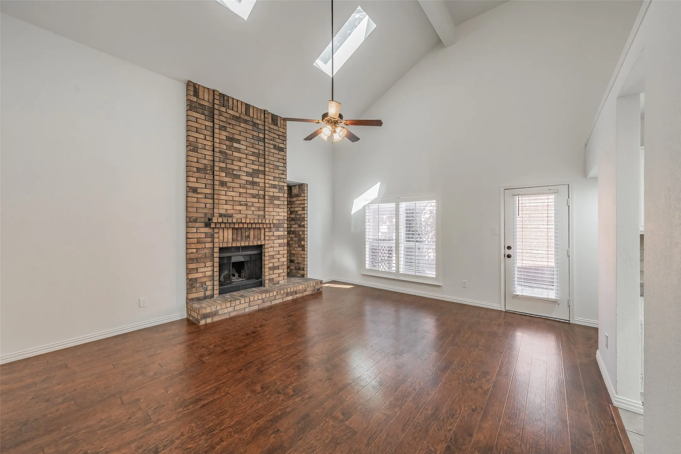 Vaulted ceiling with exposed beam, wood flooring, brick fireplace, multiple skylights, and a ceiling fan in the living room