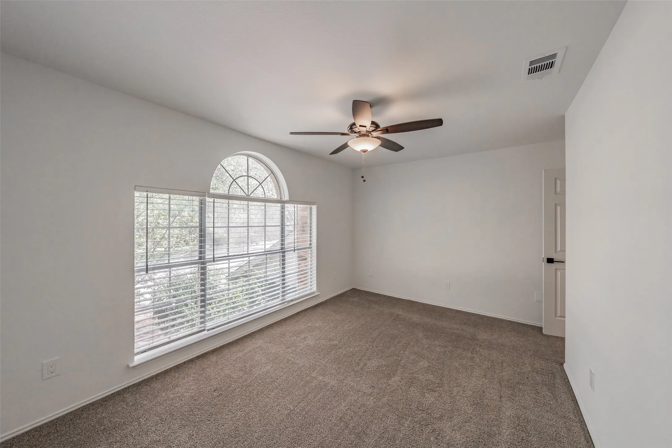 Arched transom window above a large rectangular window, textured carpeting, neutral wall paint, and a ceiling fan with light fixture