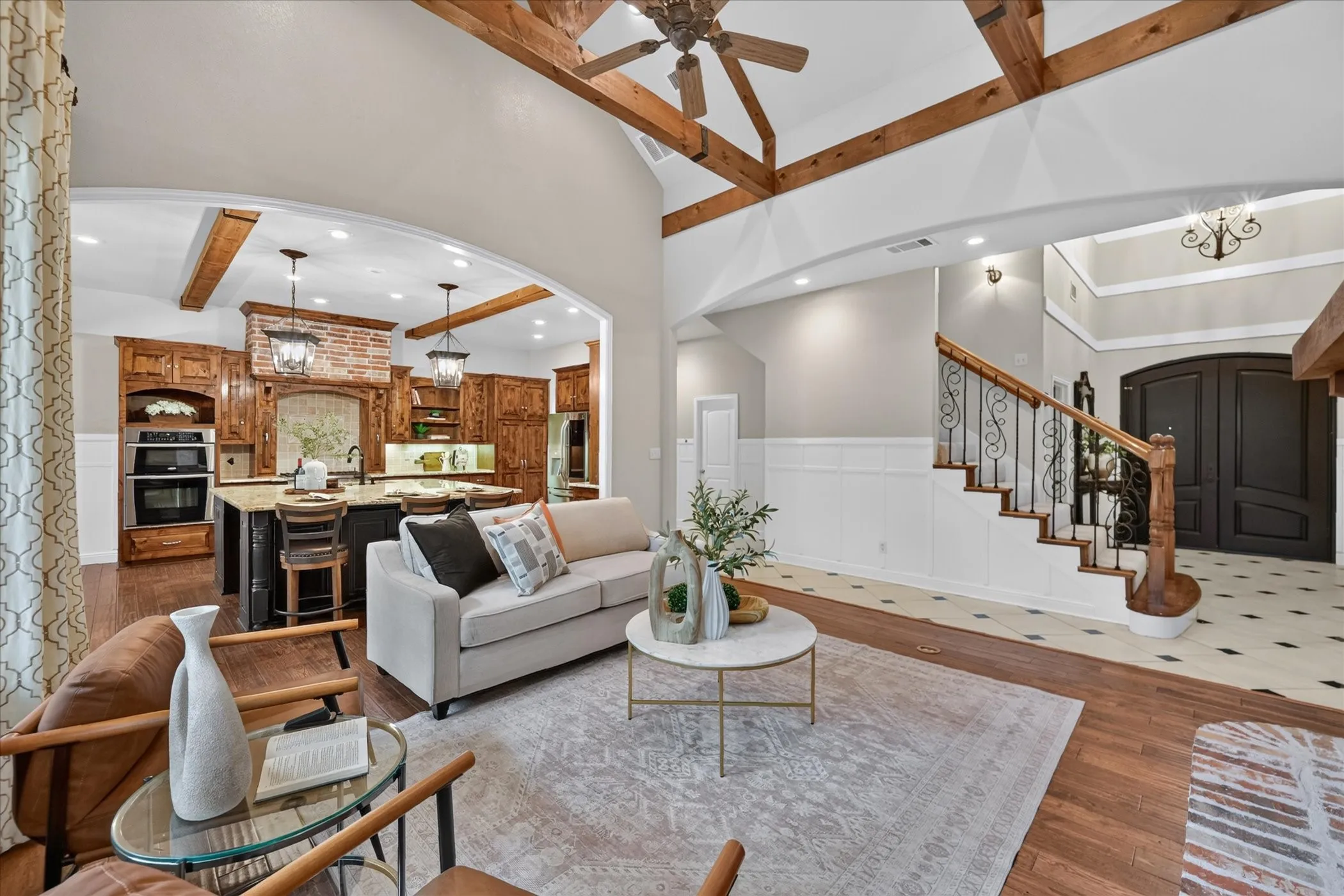 Vaulted ceiling with exposed wood beams, wood-finish flooring, staircase with ornate iron balusters and wood handrail, kitchen with double ovens and stone countertops, and a double-door entry