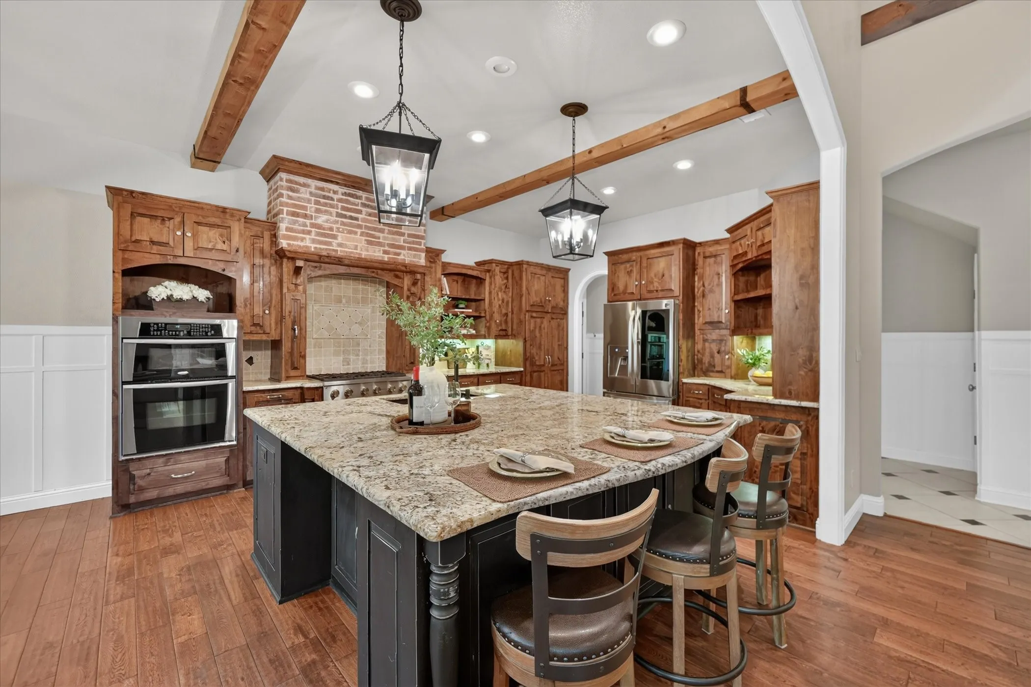 Kitchen featuring wood-finish flooring, exposed wood ceiling beams, a large island with a stone countertop, built-in stainless steel double ovens, and custom wood cabinetry