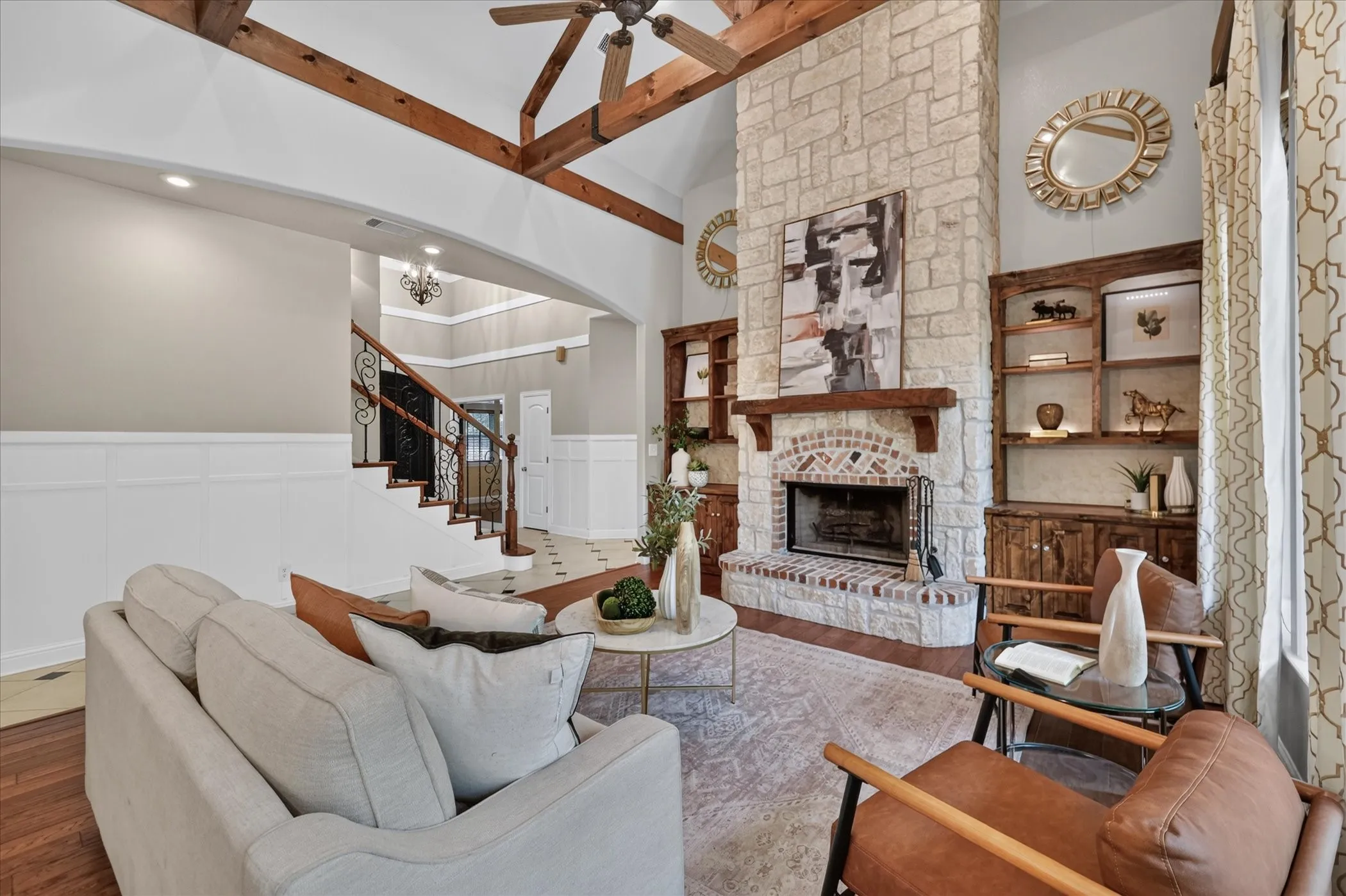 Vaulted wood-beam ceiling with a ceiling fan, stone fireplace with a wood mantle, built-in shelving with lower cabinetry, wainscoting, and wood-finish flooring