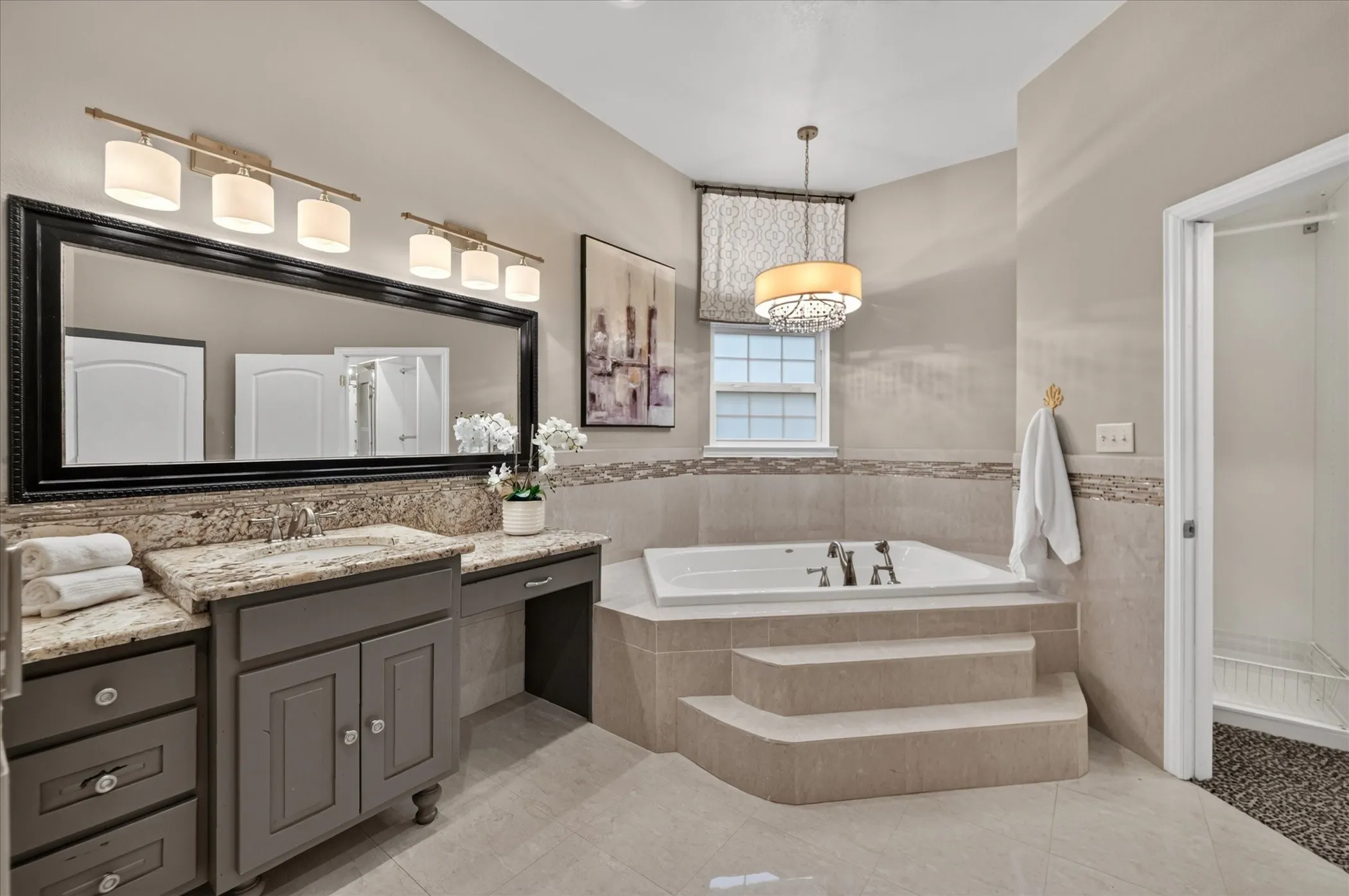 Built-in soaking tub with tiled surround, dual vanity with granite countertop, large framed mirror, and pendant light fixture
