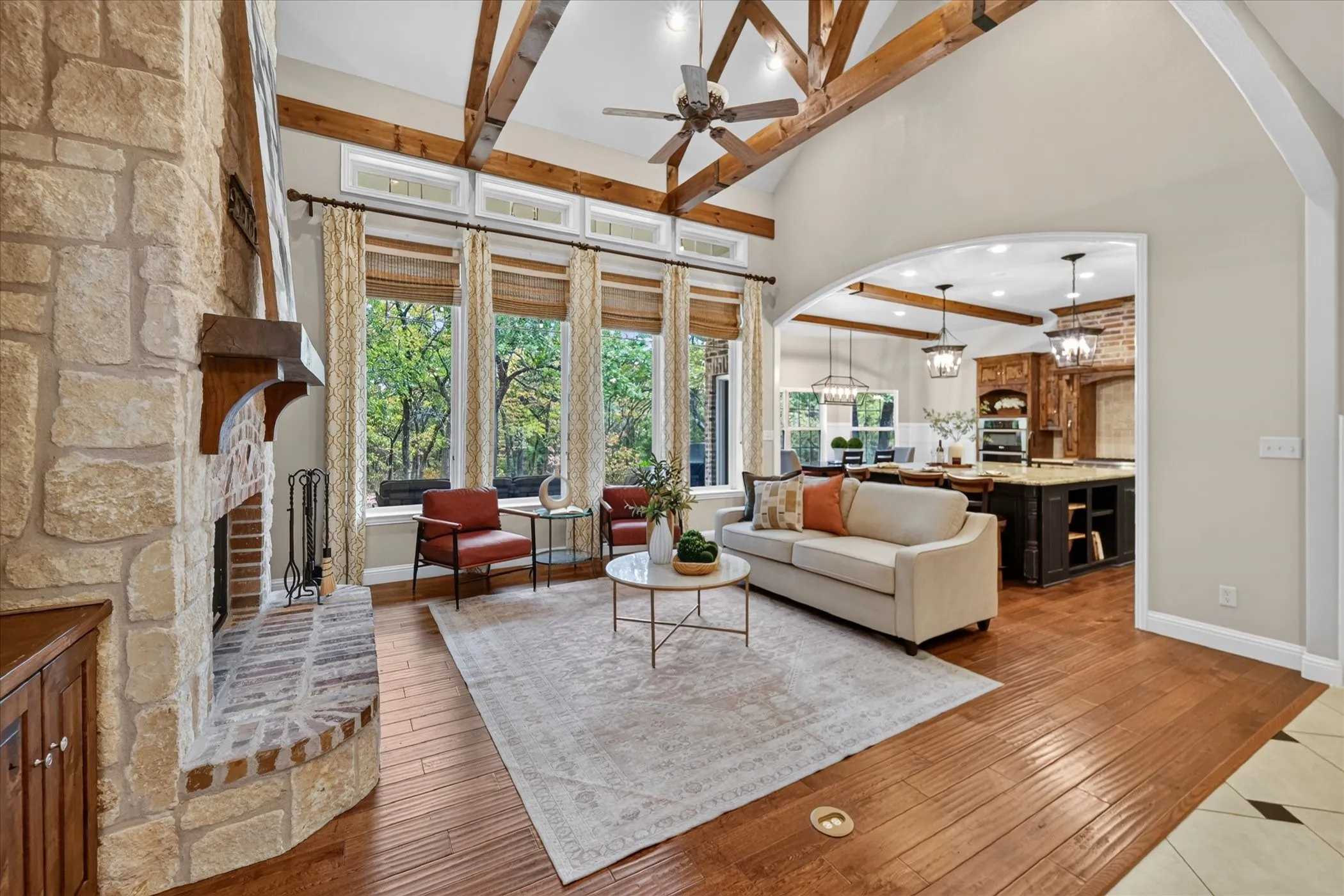 Vaulted ceilings with exposed beams, stone fireplace with a wood mantle, large floor-to-ceiling windows, wood-finish flooring, and an arched doorway