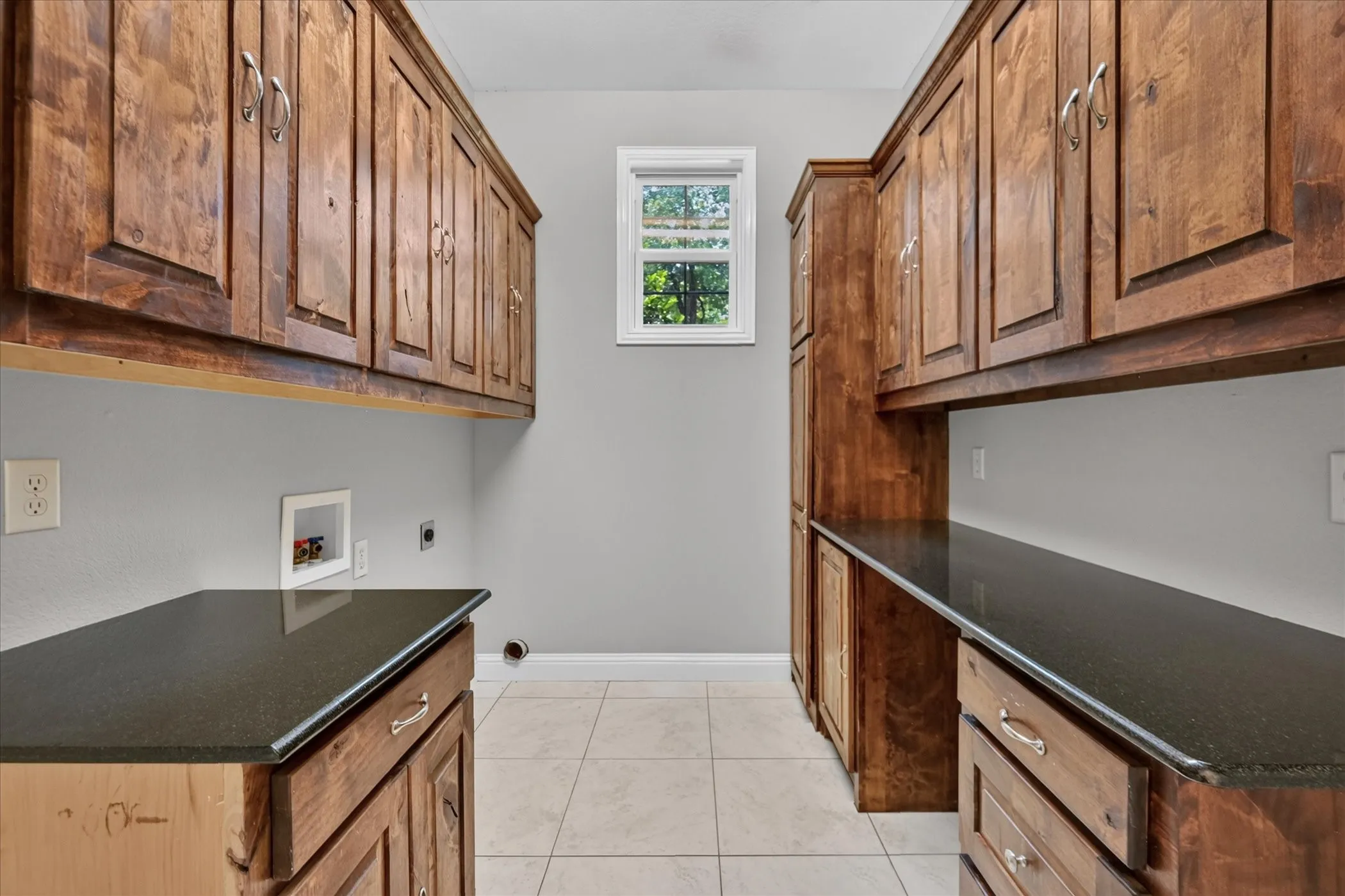 Wood-finish cabinetry with silver hardware, dark countertops, a single window, and light-toned tile flooring