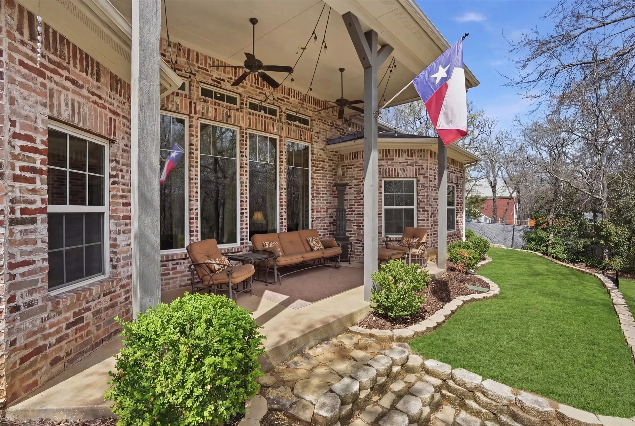 Covered patio with brick columns, ceiling fans, string lighting, and extensive window-lined walls overlooking a manicured lawn
