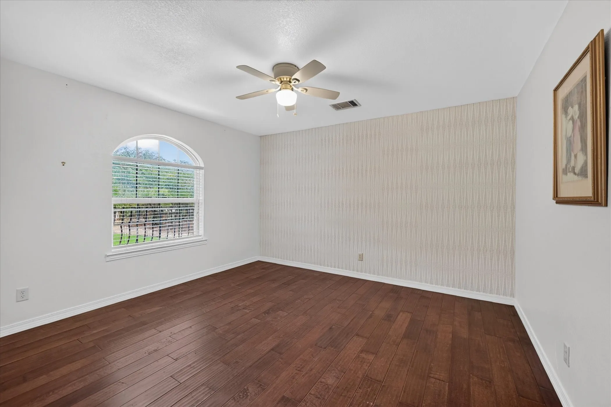Wood-finish flooring, arched window with horizontal blinds, ceiling fan with integrated lighting, and a textured accent wall