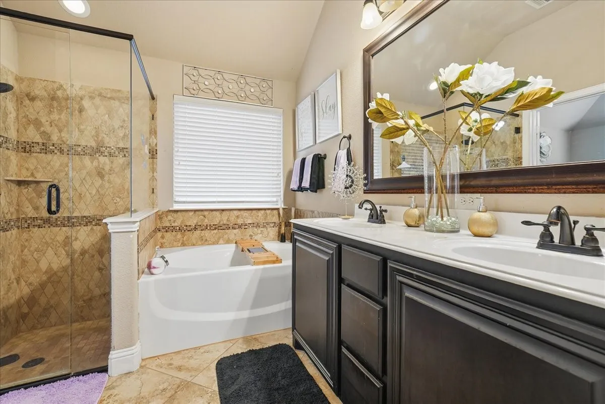 Walk-in shower with natural stone tile, garden tub, double vanity with dark wood-finish cabinetry, and a large framed mirror