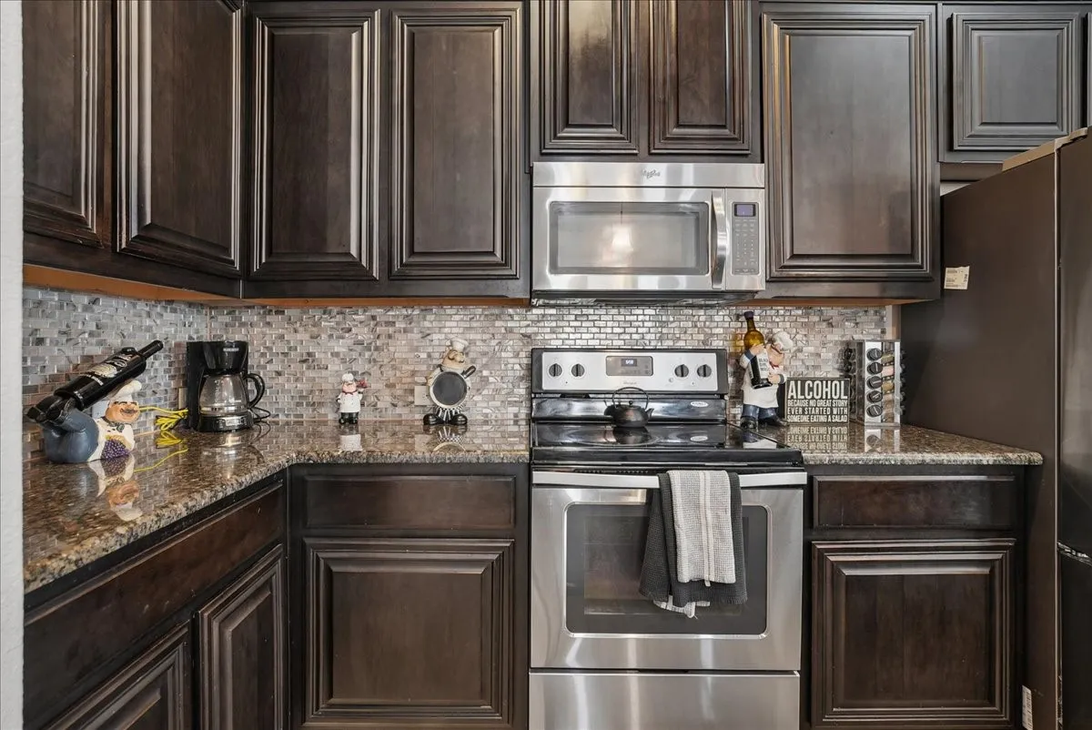 Kitchen featuring dark wood cabinetry, a mosaic tile backsplash, polished stone countertops, and stainless steel appliances including a microwave and range