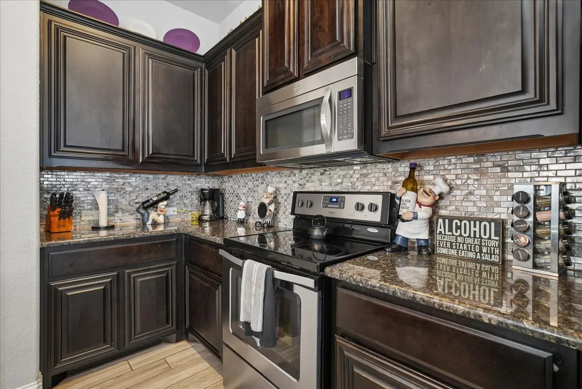 Dark wood cabinetry, stainless steel appliances, mosaic tile backsplash, granite countertops, and wood-finish flooring