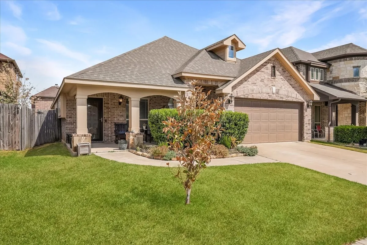 Brick facade home featuring a covered front porch, an attached garage, a dormer window, and mature landscaping