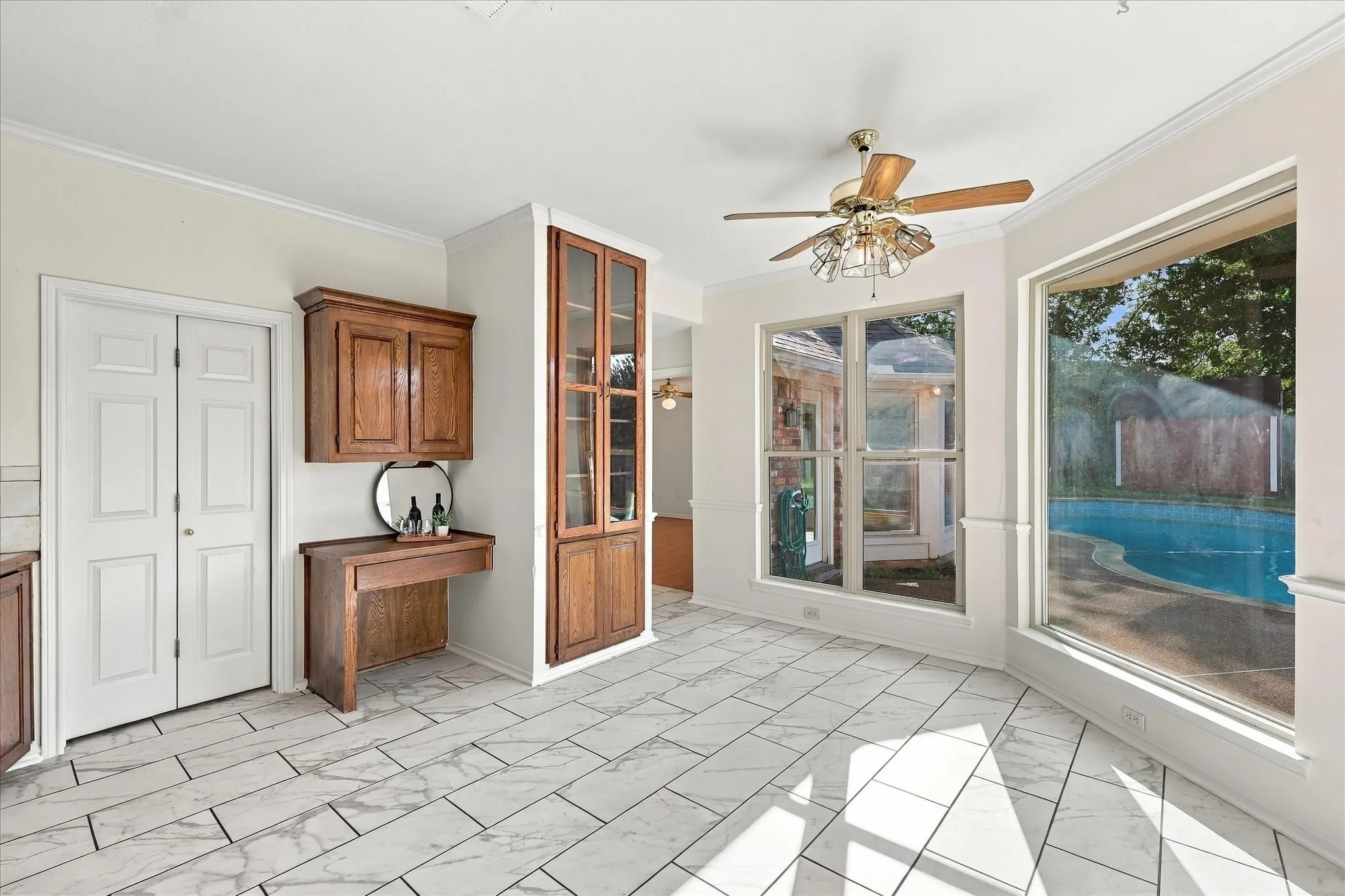 Light-toned tile flooring, built-in wooden cabinetry, ceiling fan, and large windows with a pool visible
