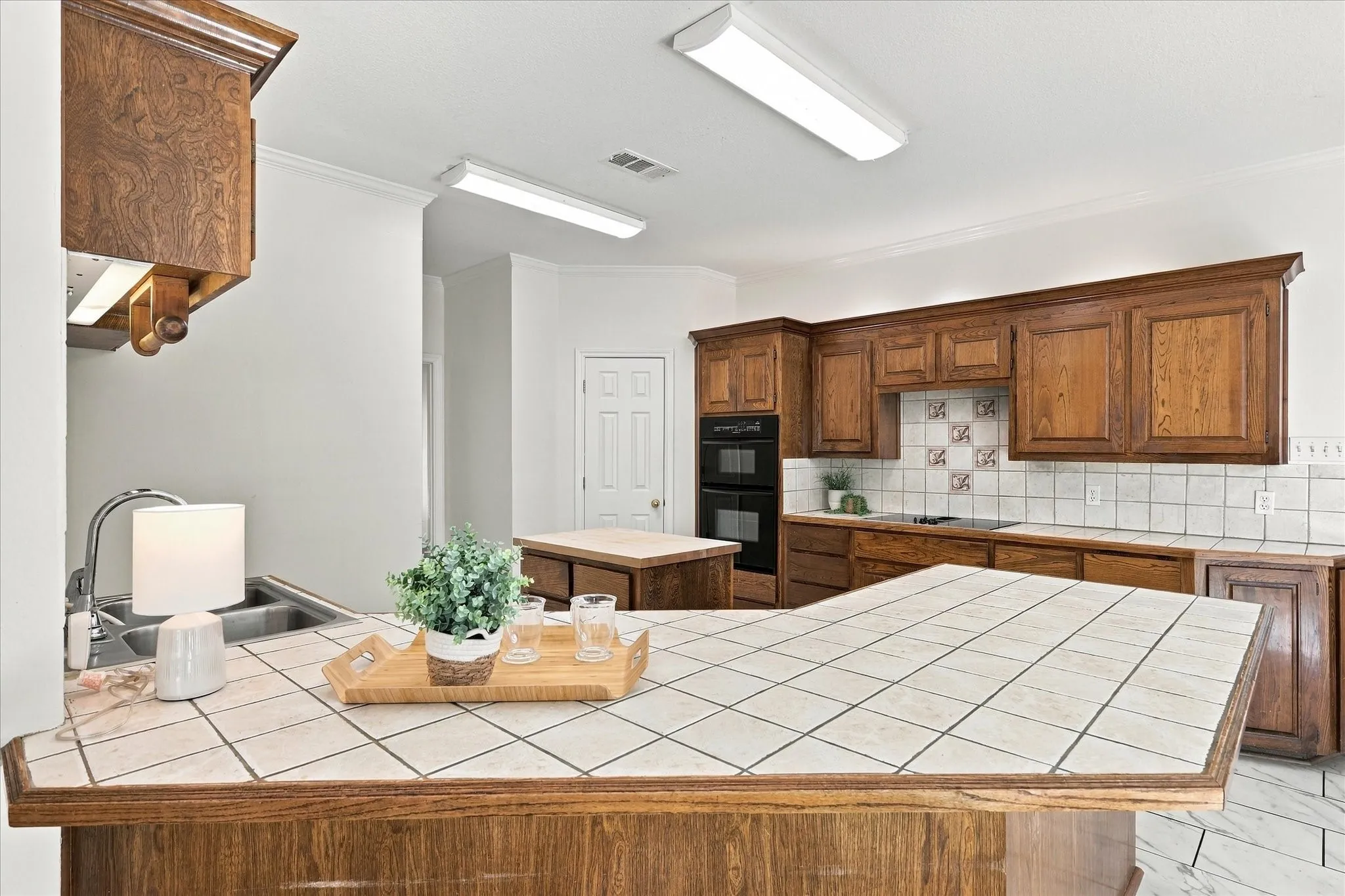 Kitchen with wood-finish cabinetry, tile countertops, tile backsplash, stainless steel sink, and built-in microwave and oven