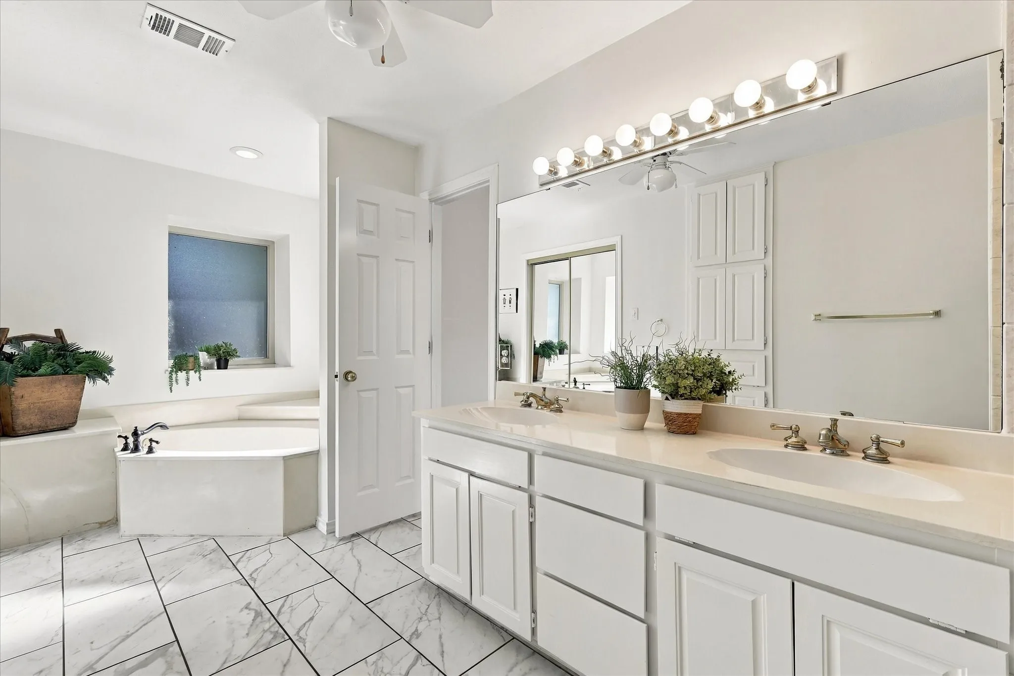 Dual vanity with undermount sinks and brushed brass fixtures, large wall-mounted mirror, built-in storage cabinetry, corner soaking tub with deck-mounted faucet, and tile flooring