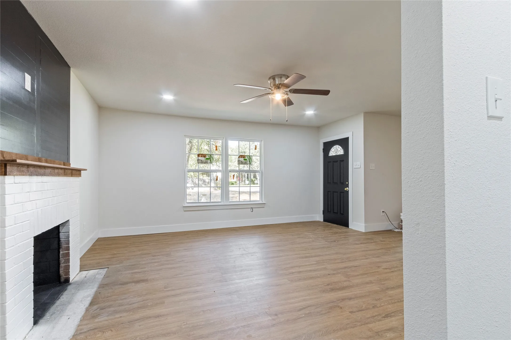 Wood-finish flooring with white baseboards, a white brick fireplace with a wood mantle, a ceiling fan with integrated lighting, recessed ceiling lights, and a dark entry door with decorative glass