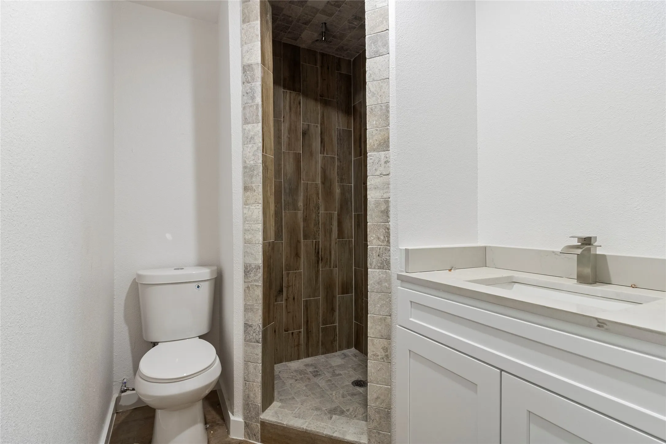 Shower with wood-finish tile surround, stone tile shower floor, single vanity with integrated sink, stone countertop, and brushed nickel faucet