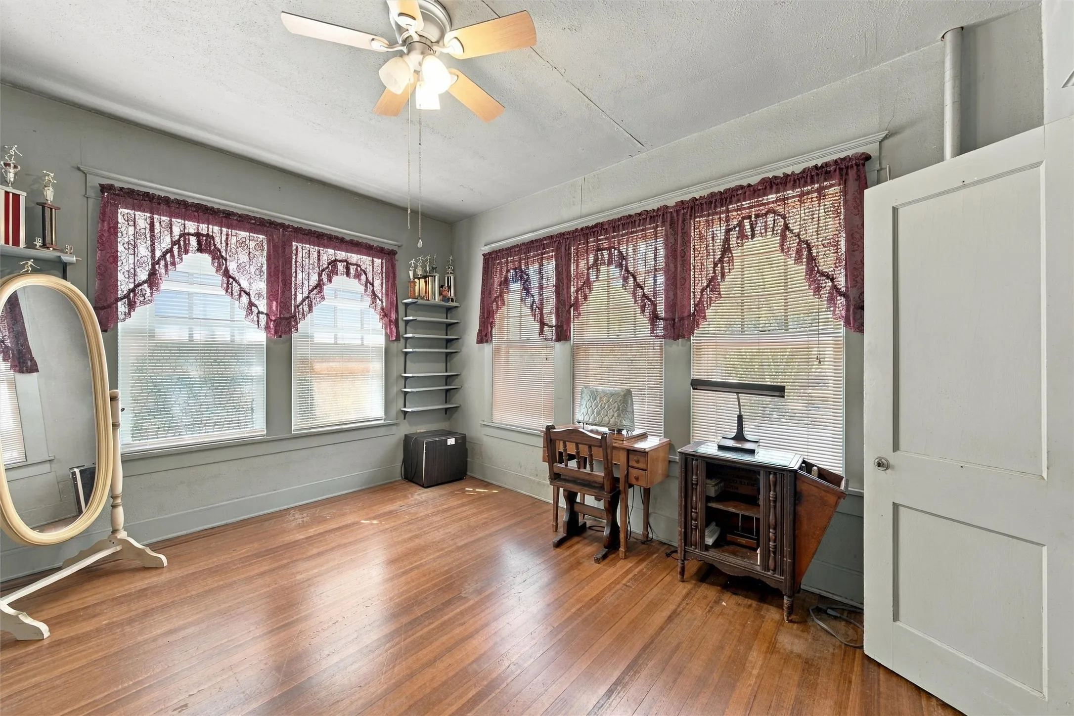 Spacious room featuring hardwood flooring, ceiling fan with light fixture, and multiple windows with burgundy swag valances