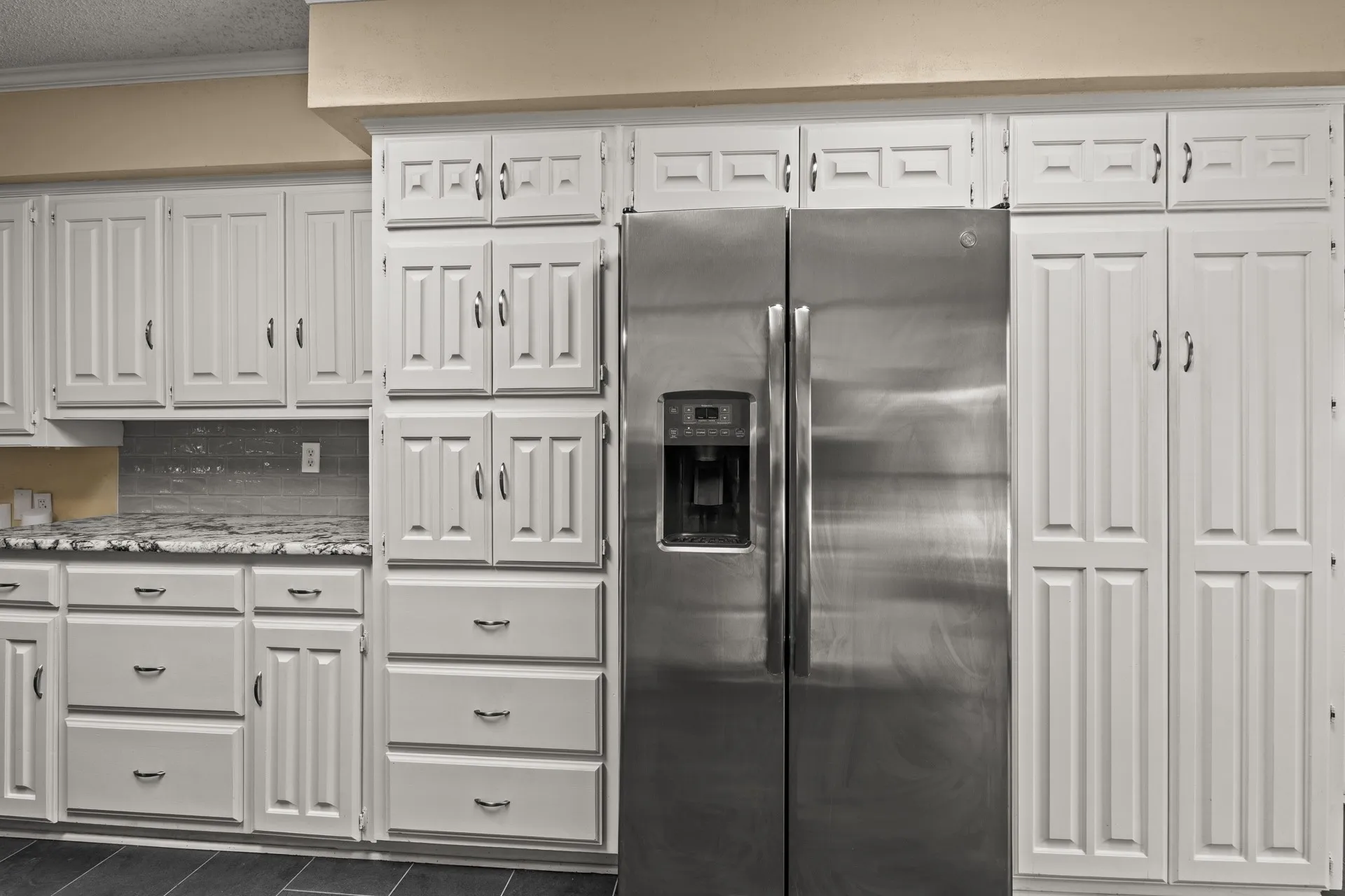 Kitchen with stainless steel fridge, ornamental molding, white cabinetry, dark tile patterned flooring, and light stone counters