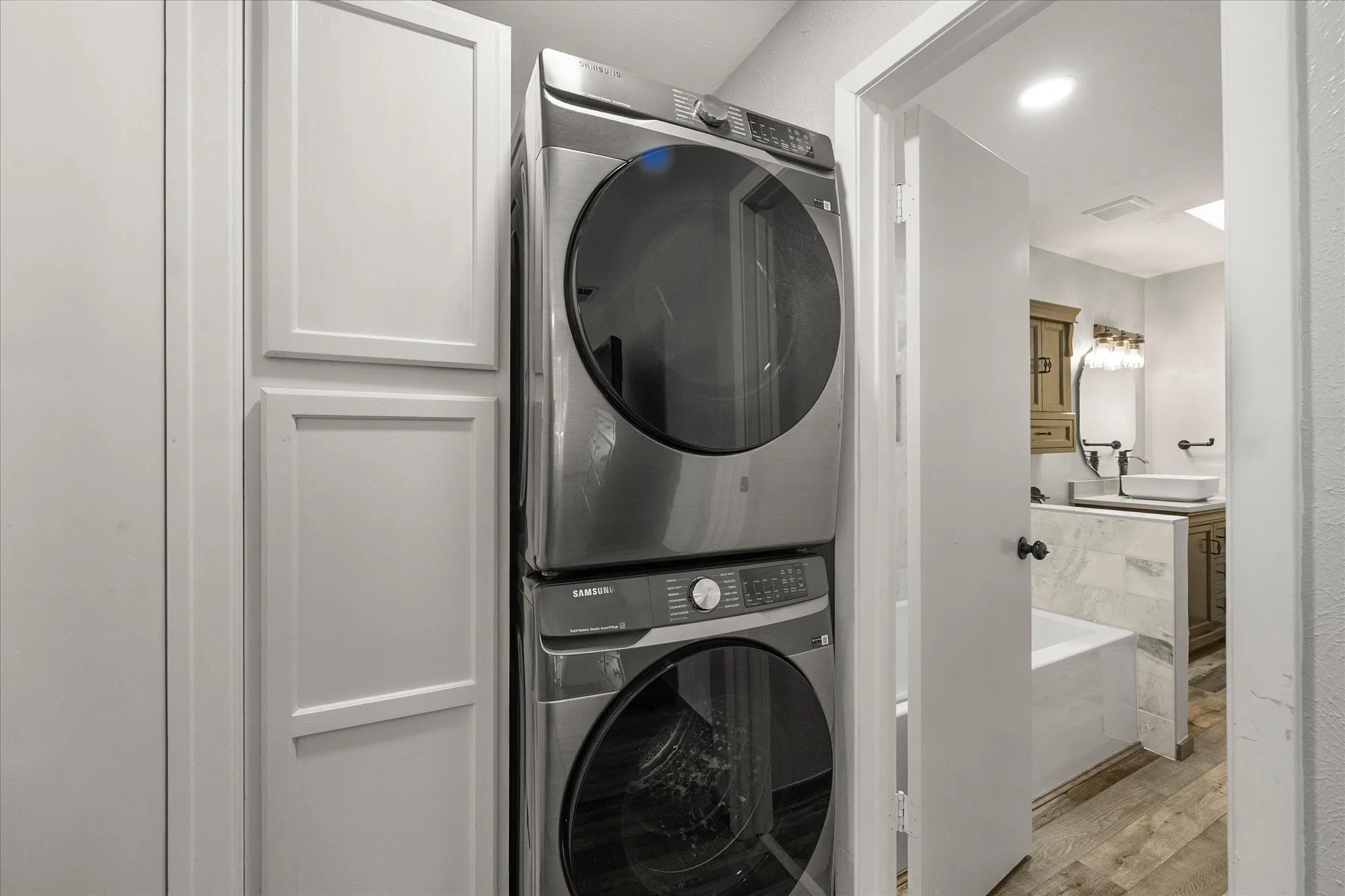 Built-in white cabinetry, stackable washer and dryer, wood-finish vinyl plank flooring, white soaking tub, and a vanity with a vessel sink