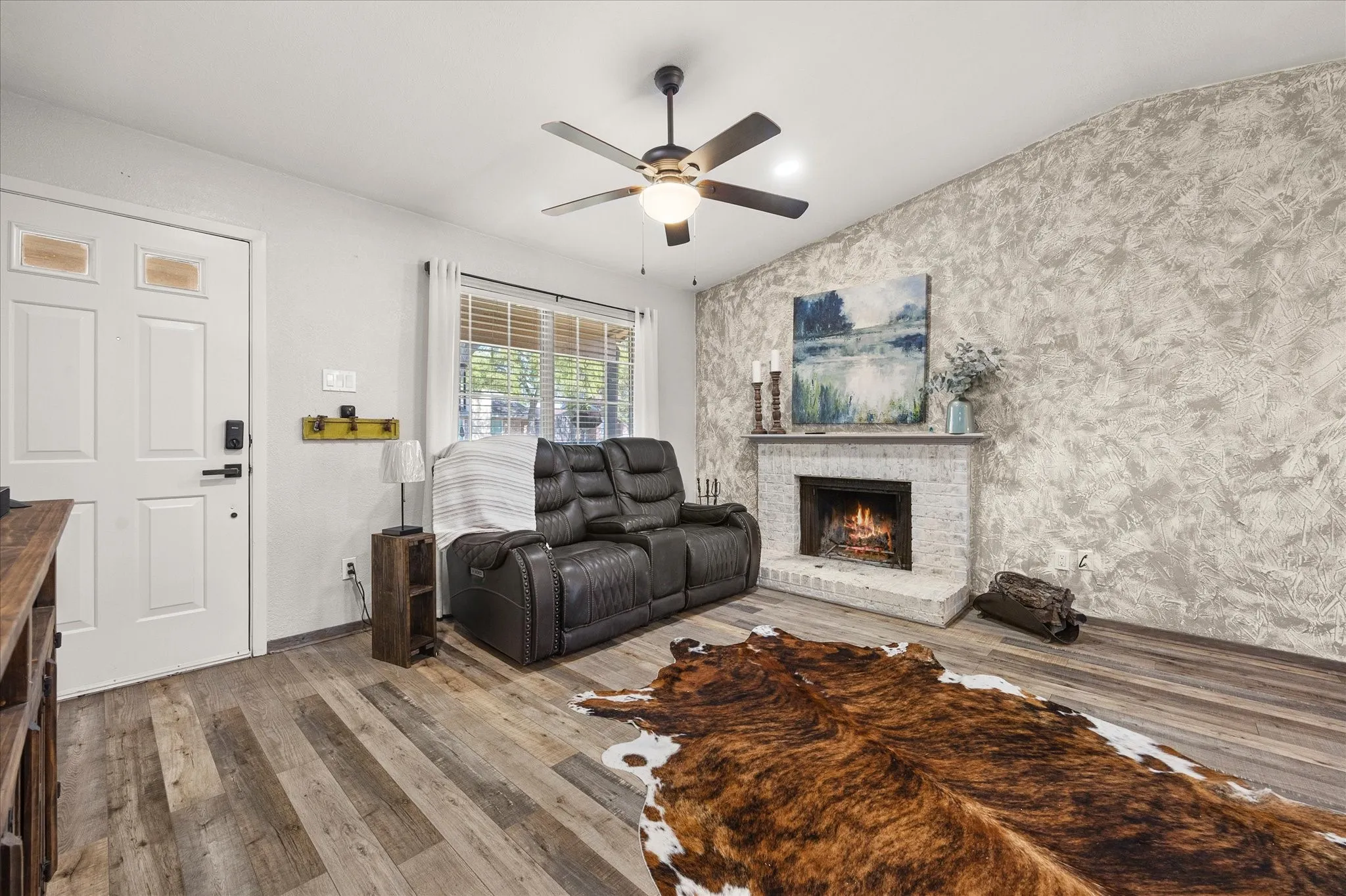 Brick fireplace, wood-finish vinyl plank flooring, ceiling fan, white paneled door, and window with blinds