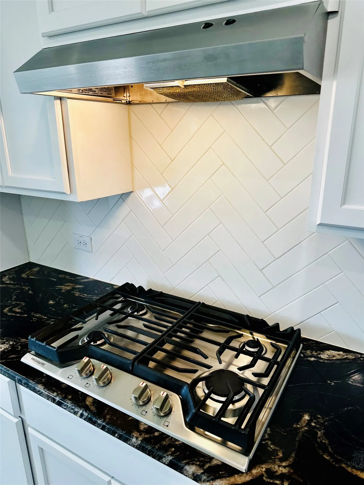 Stainless steel range hood with integrated lighting over a gas cooktop, set against a white subway tile backsplash in a herringbone pattern
