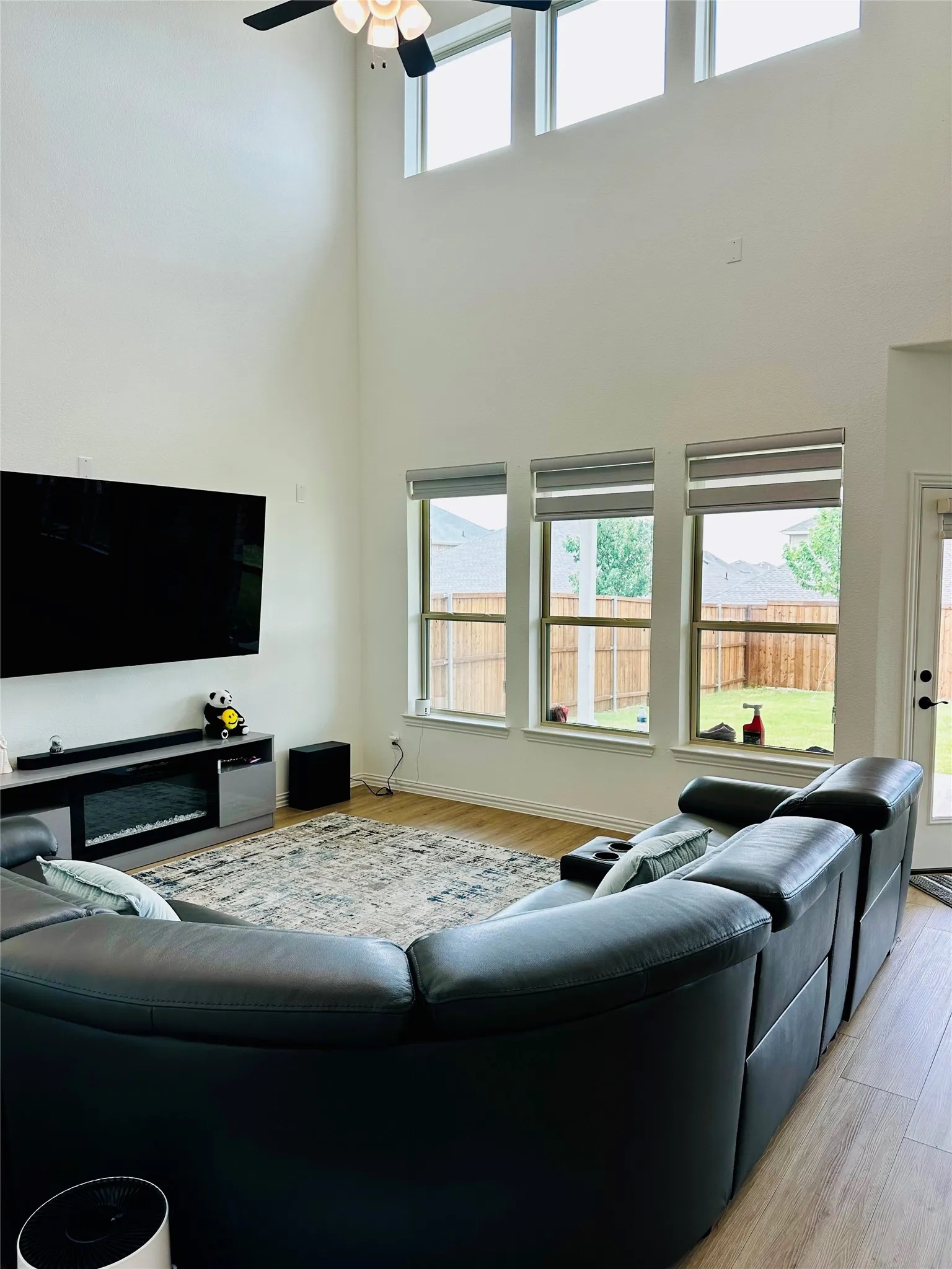 Soaring ceiling with high windows, three lower windows with roller shades, wood-finish flooring, and a built-in electric fireplace
