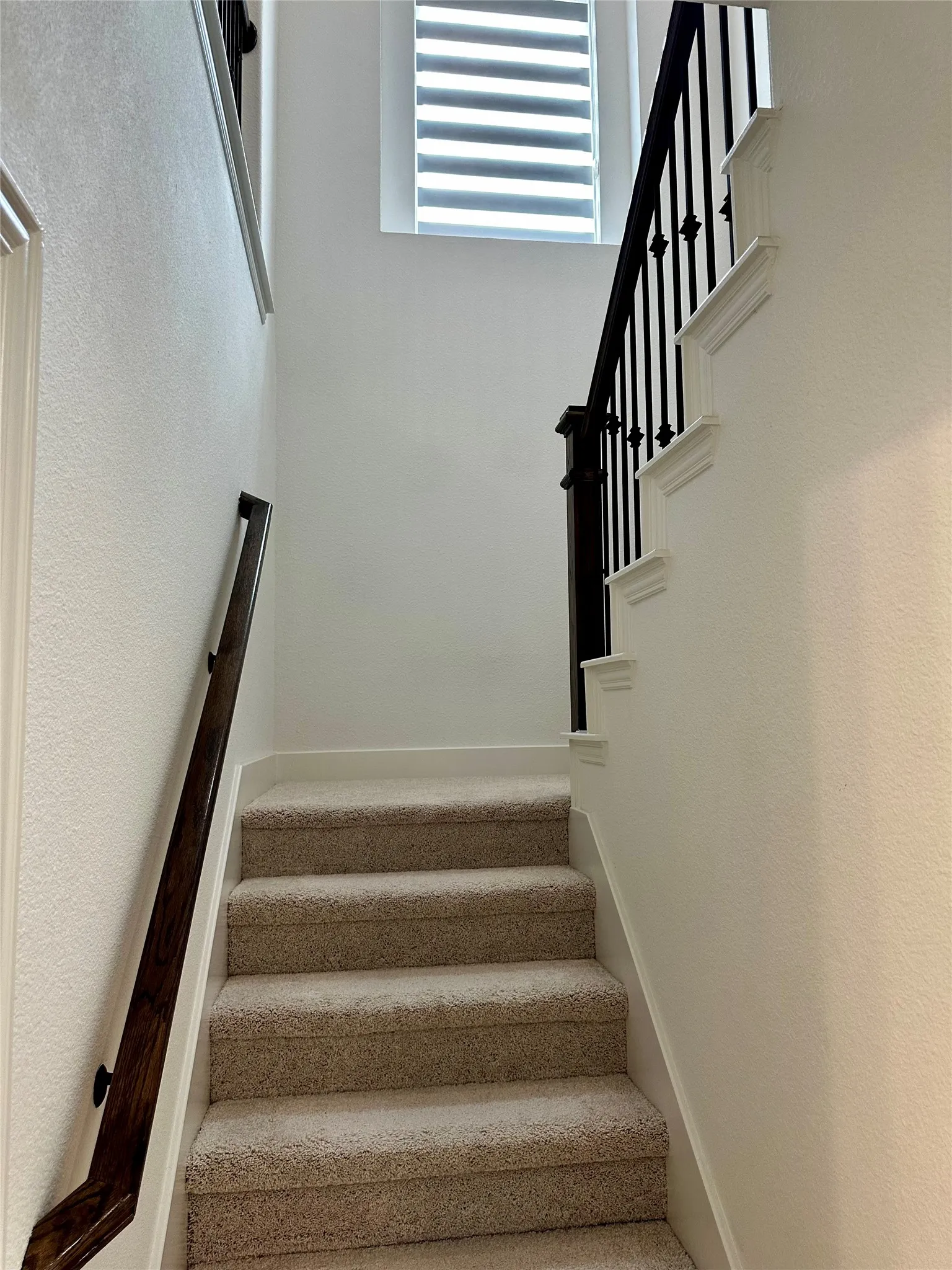 Carpeted staircase with a dark wood handrail, black wrought iron balusters, and a window with horizontal blinds