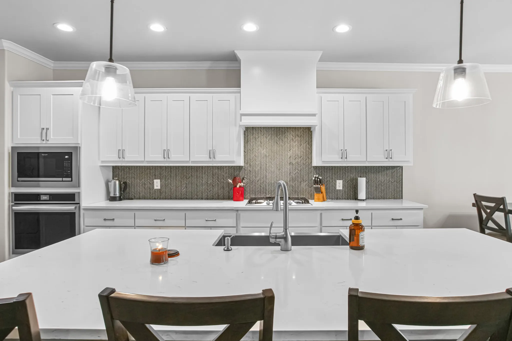 White shaker cabinetry, built-in microwave and oven, island with integrated sink, patterned tile backsplash, and recessed and pendant lighting