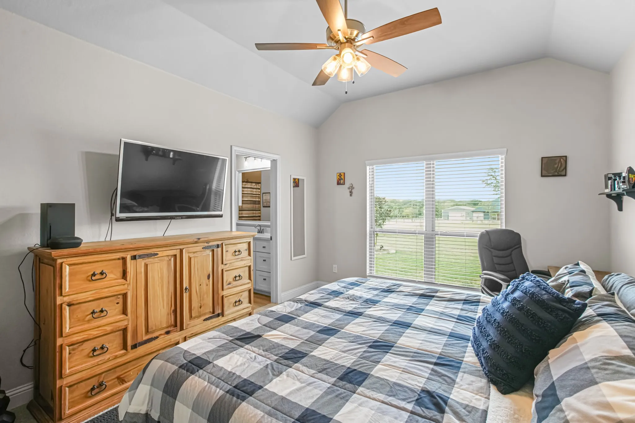 Vaulted ceiling with ceiling fan, large window with blinds, and an attached bathroom with a vanity and mirror