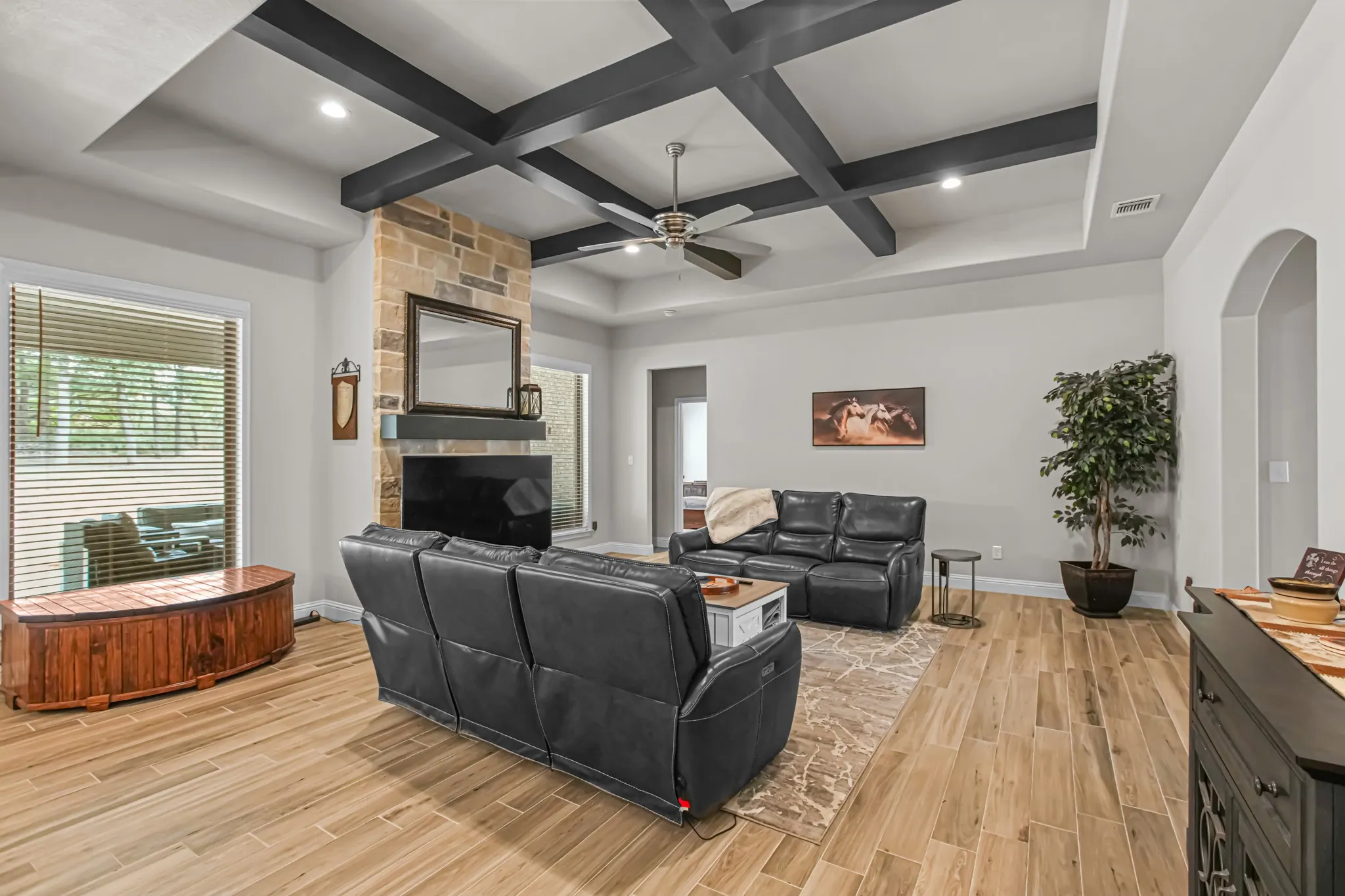 Coffered ceiling with recessed lighting and a ceiling fan, stone-clad fireplace with a wood mantel, arched entryway, and wood-finish flooring