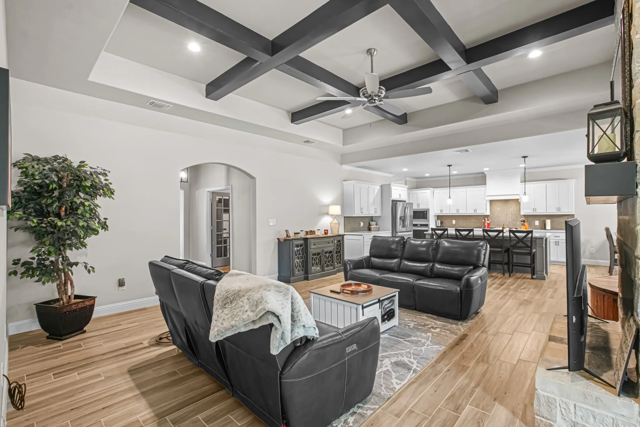 Coffered ceiling with a ceiling fan, light wood-finish flooring, stone fireplace, arched doorway, and recessed lighting