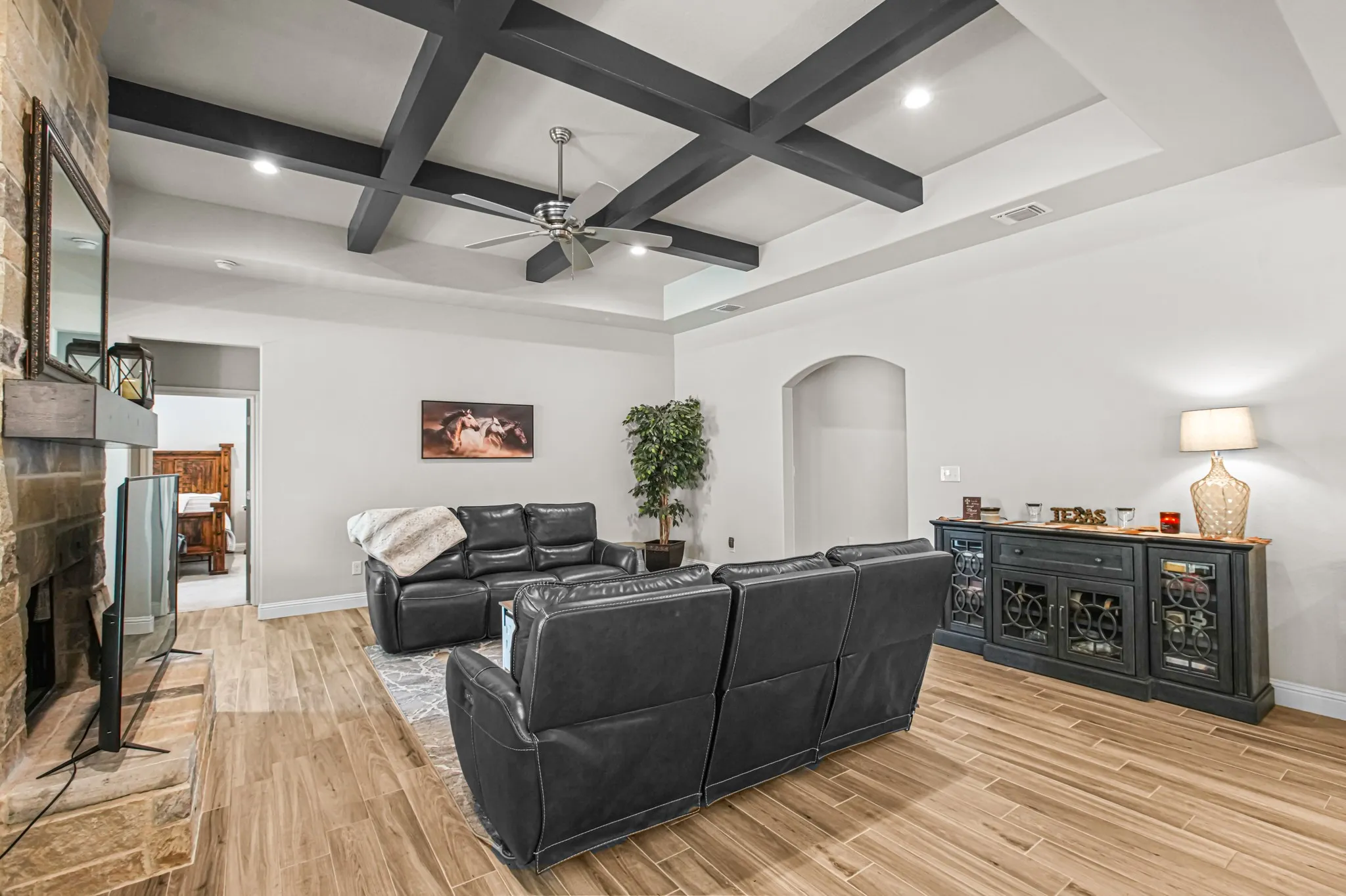 Coffered ceiling with recessed lighting, stone-clad fireplace with wood mantle, wood-finish flooring, and an arched doorway