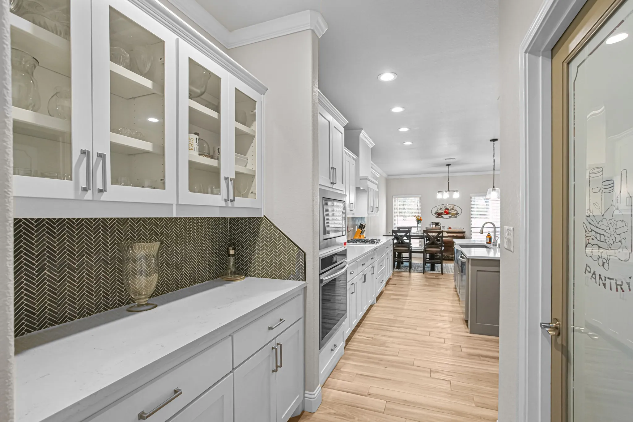 White shaker cabinetry with glass-front uppers, herringbone tile backsplash, integrated stainless steel appliances, and light wood-finish flooring